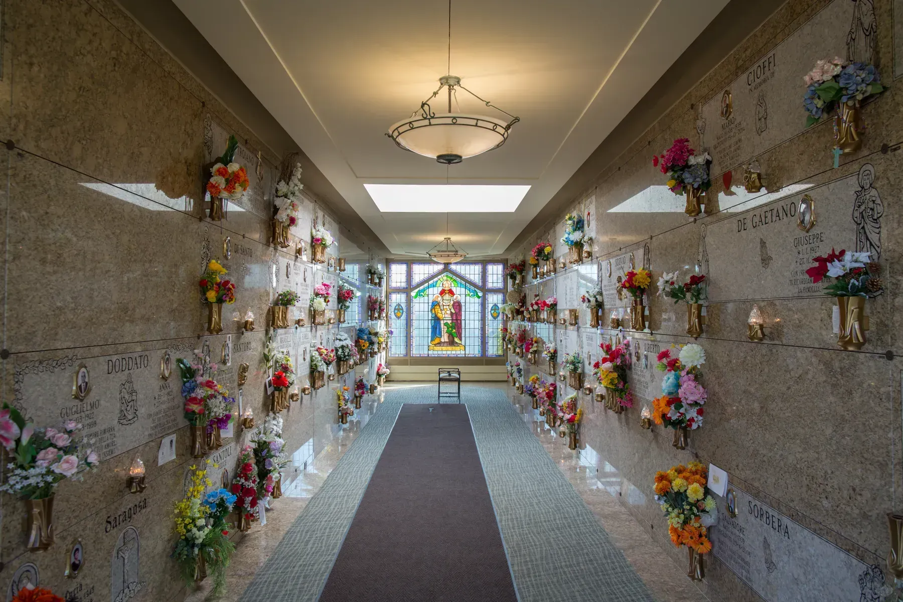 Narrow memorial hallway with flower arrangements along both walls leading to a stained-glass window.