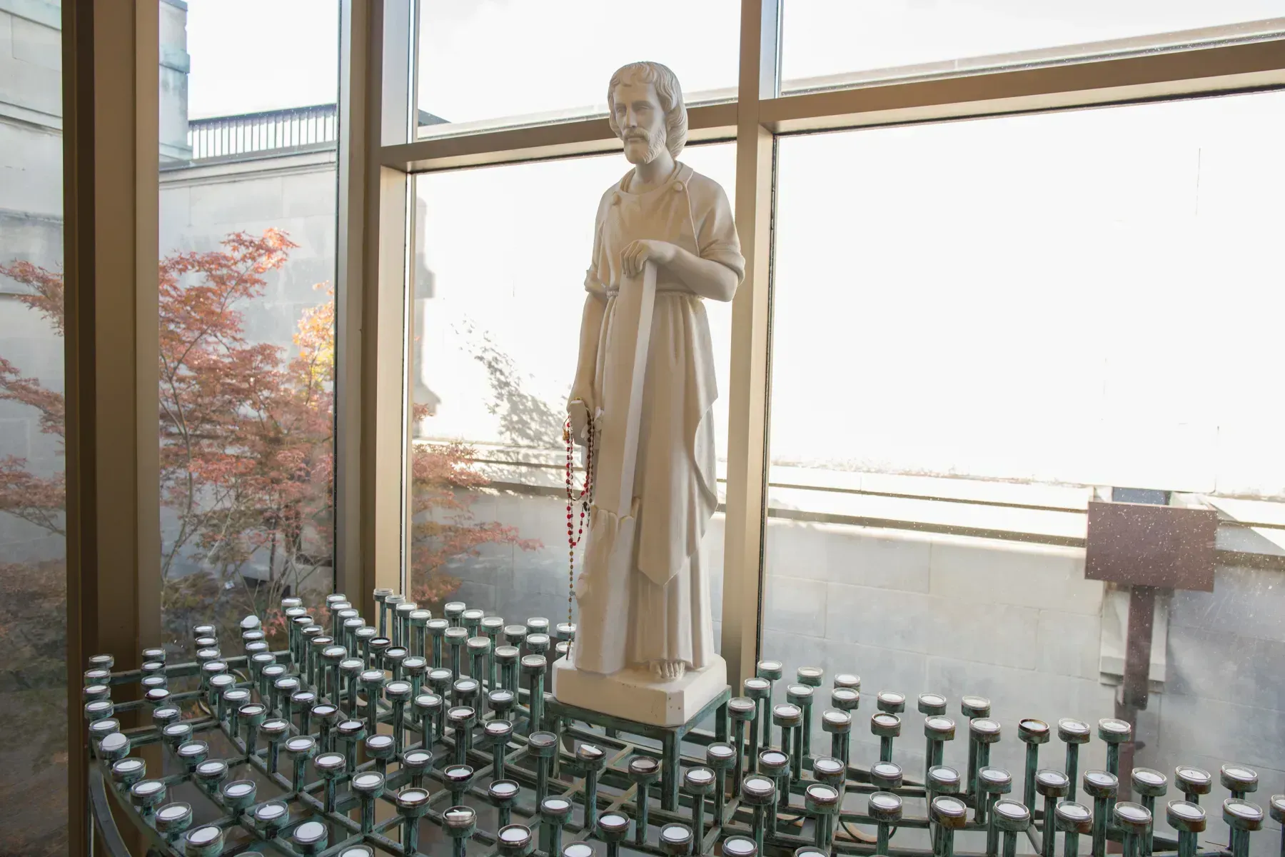 Statue of a robed figure inside a chapel, surrounded by rows of lit votive candles near tall windows