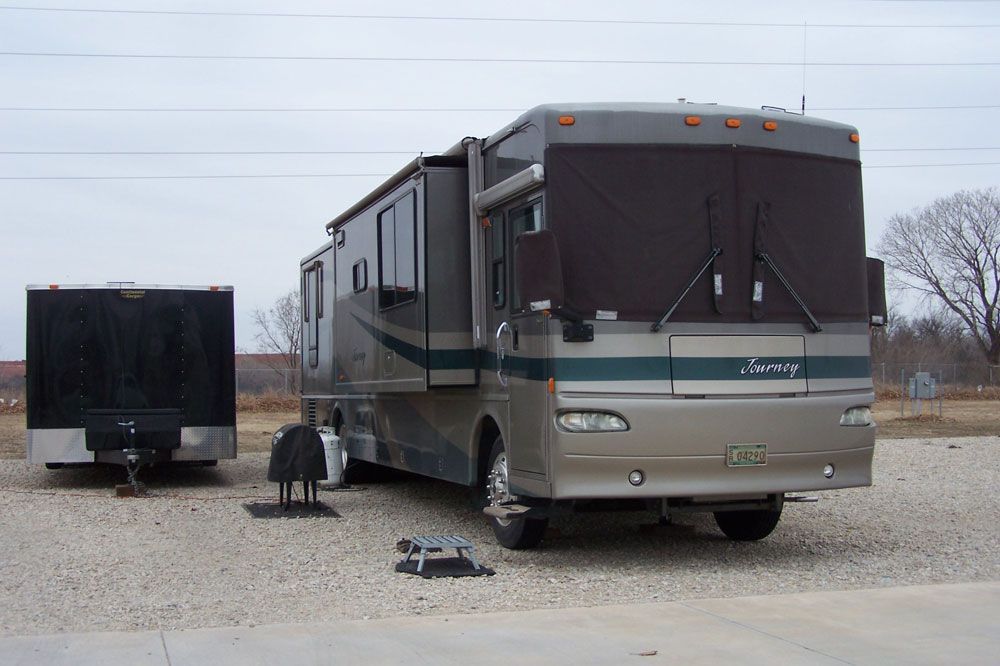 A large rv is parked in a gravel lot next to a trailer.