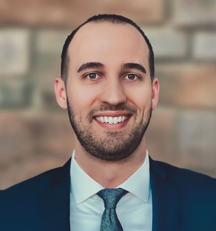 Man in a suit smiling, headshot against a brick wall background.