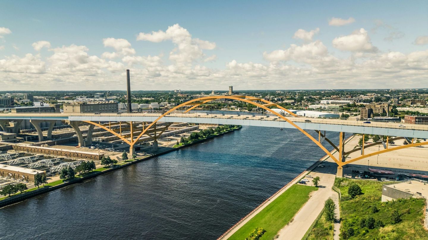 Yellow arch bridge spanning a dark river with city buildings in the background under a blue sky.