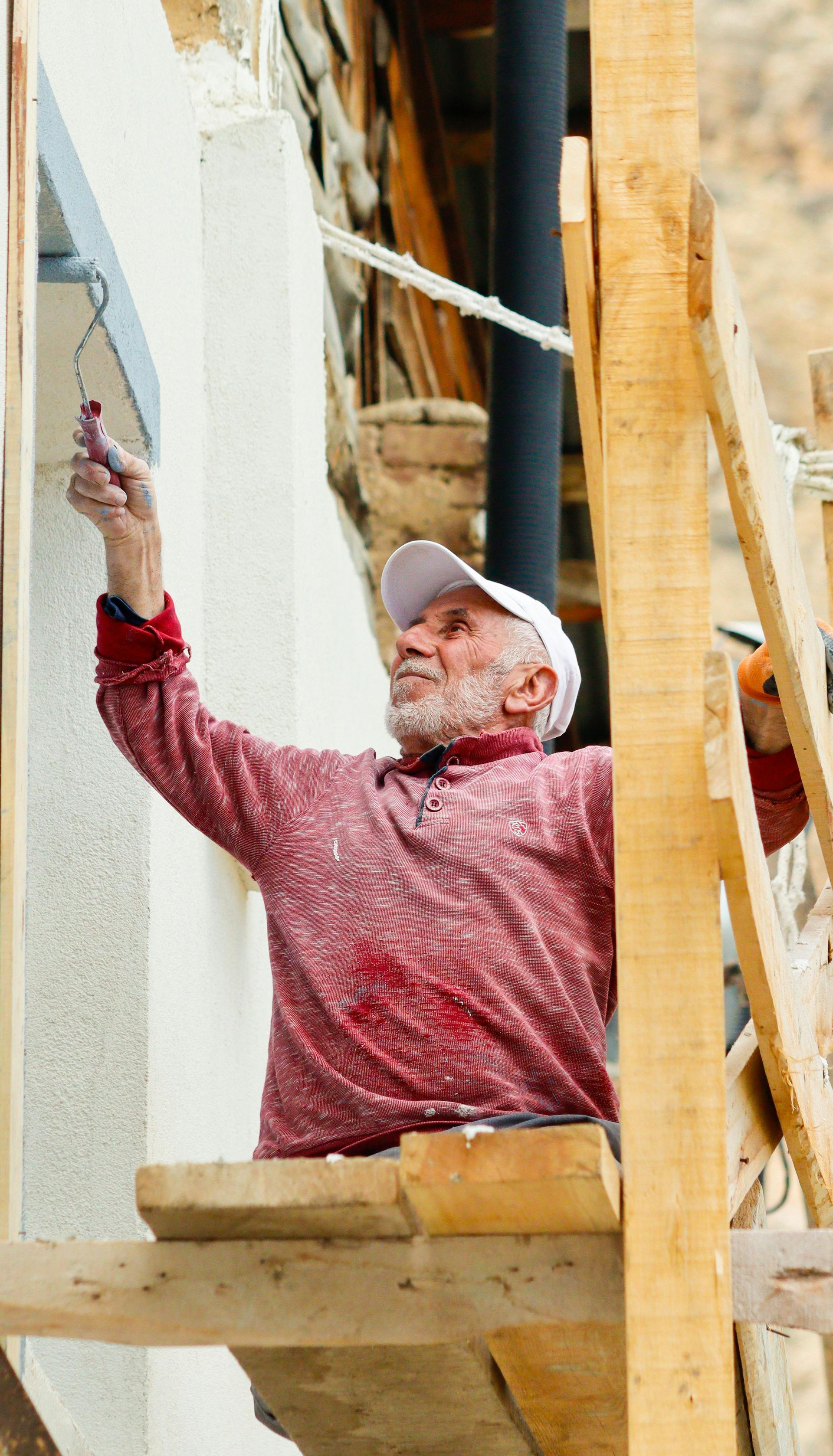 An older man is painting a wall on a scaffolding.