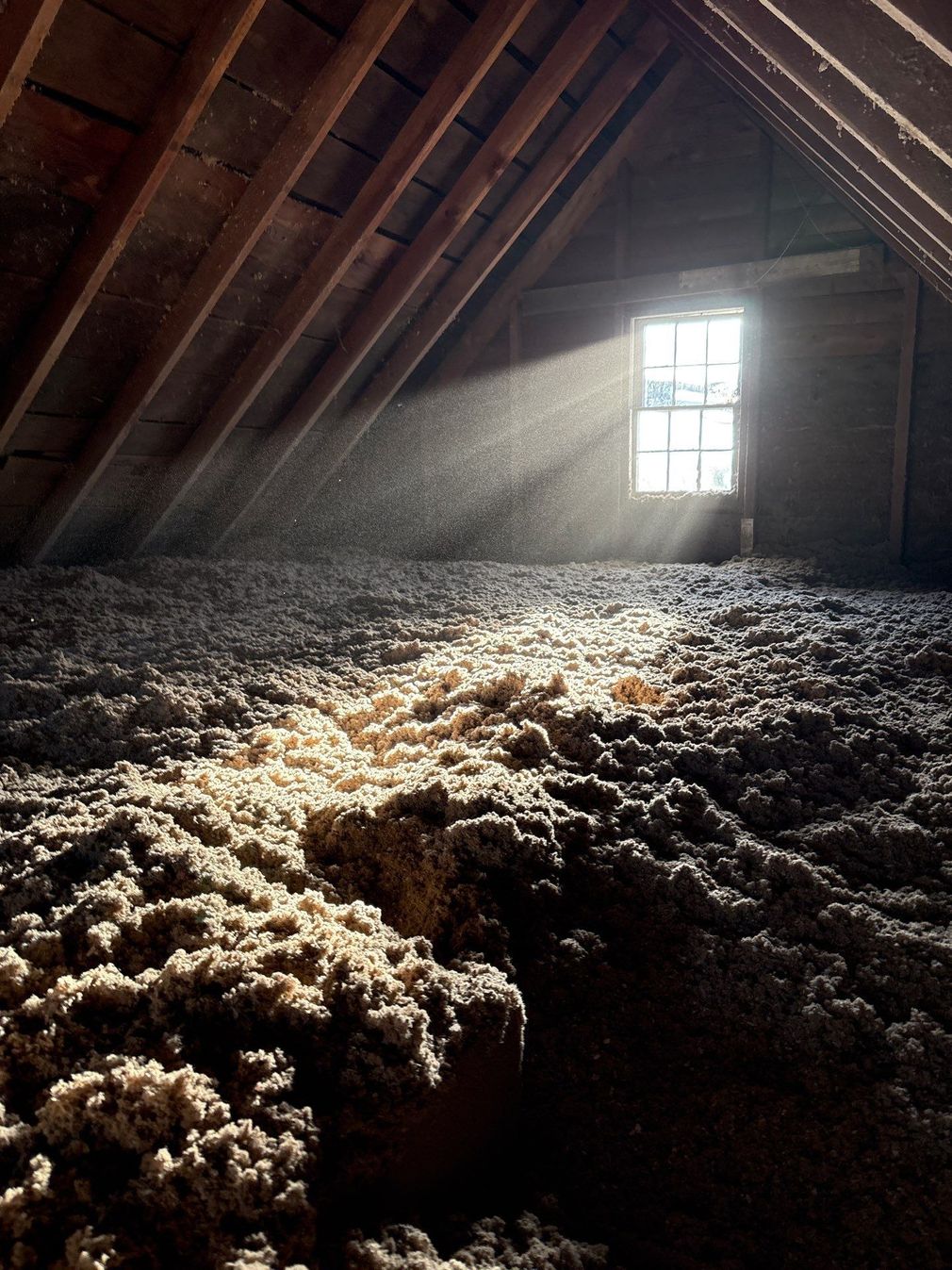 Attic with slanted wooden beams, sunbeams through a window, and piles of insulation on the floor