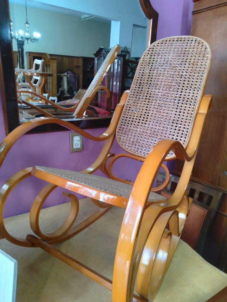 Mecedora de mimbre con brazos curvos de madera y cojín de asiento color canela en una sala de estar con espejo.