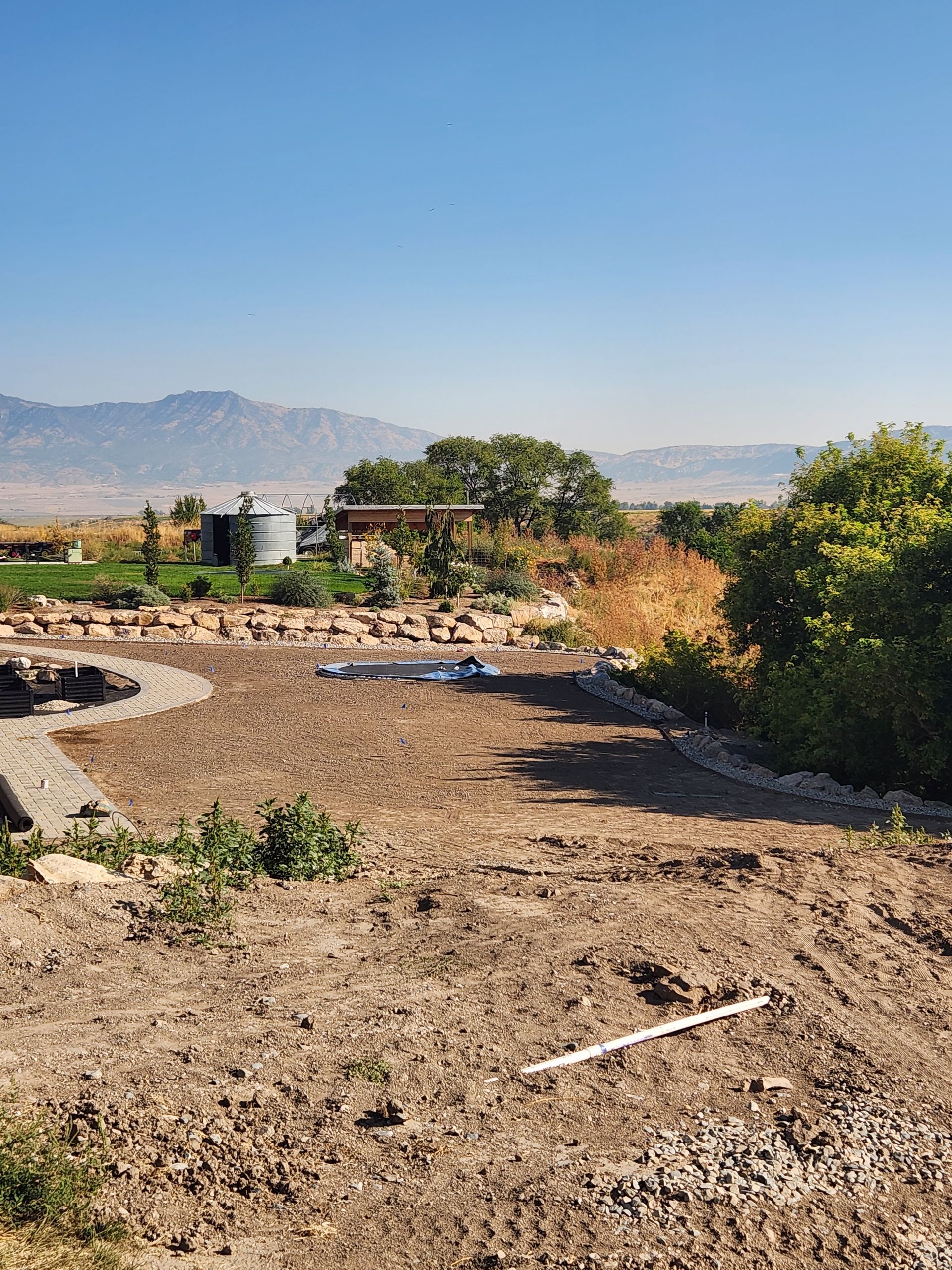 Dirt yard with a stone border, green vegetation, a silo, and mountains in the distance under a blue sky.