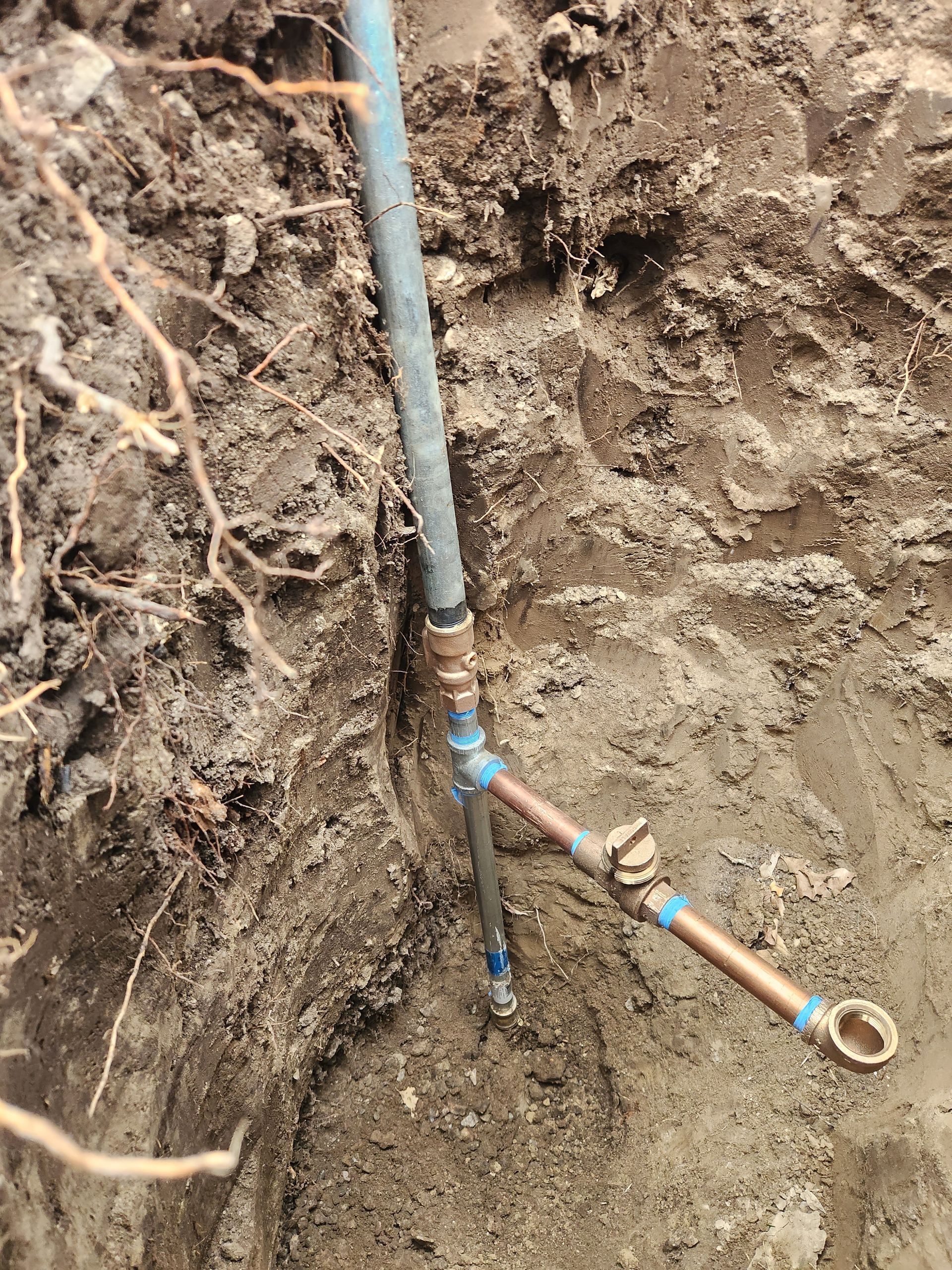 Trench dug in dirt revealing copper pipes and blue PVC pipe connected with brass fittings.