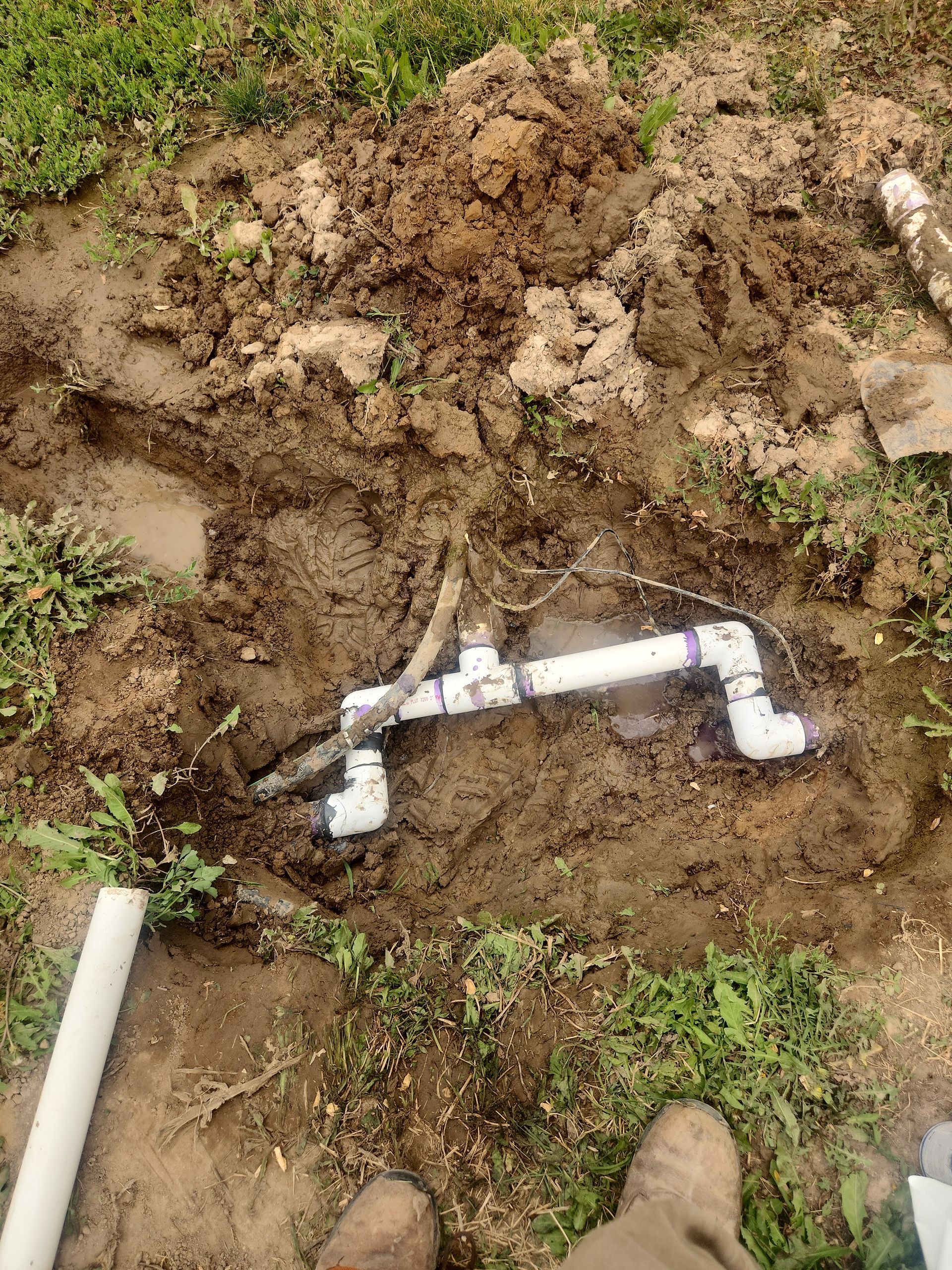 PVC pipe assembly exposed in muddy ground; lawn and grass visible.