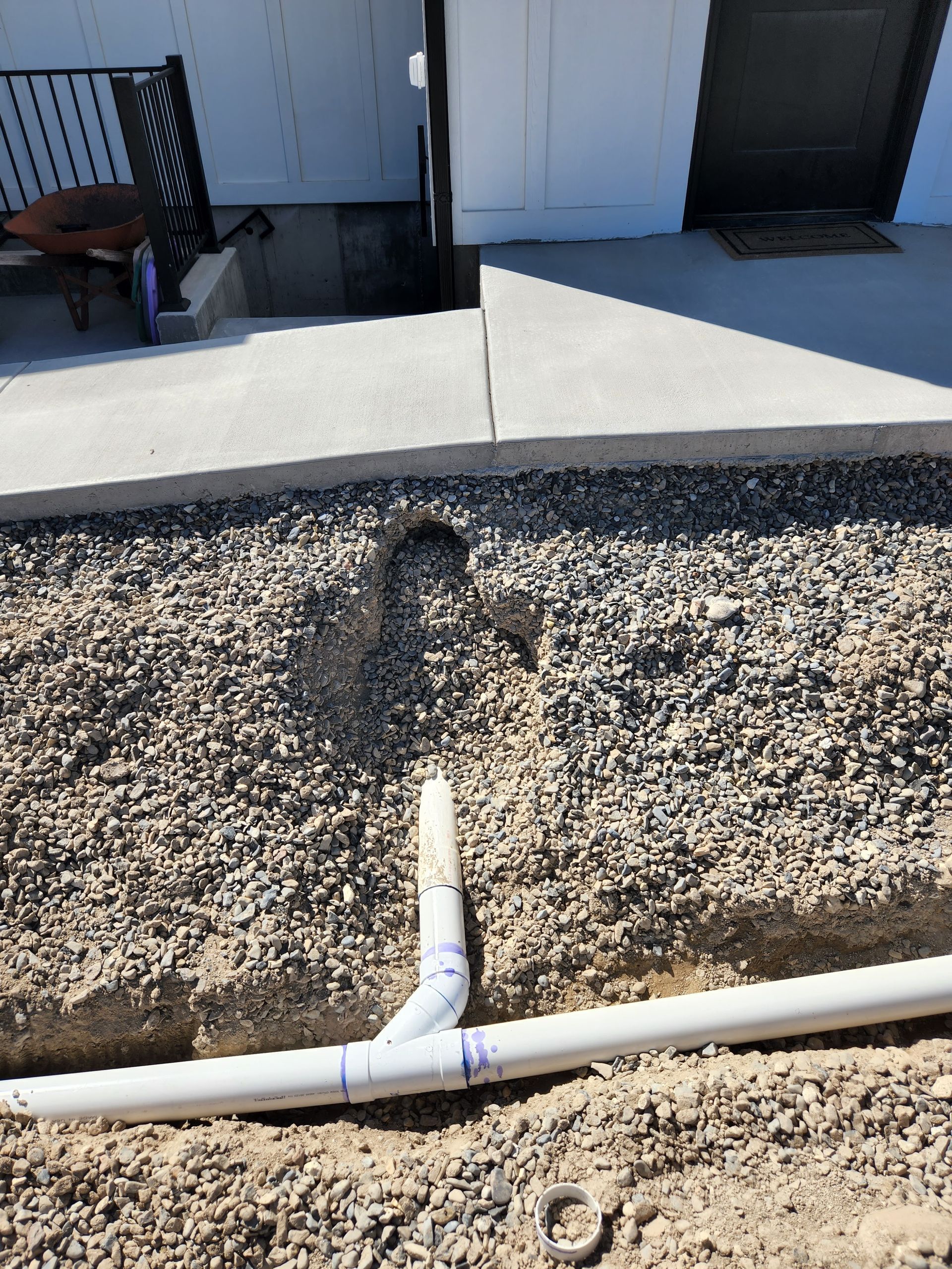 PVC pipes in a gravel trench, near a concrete walkway and white building.