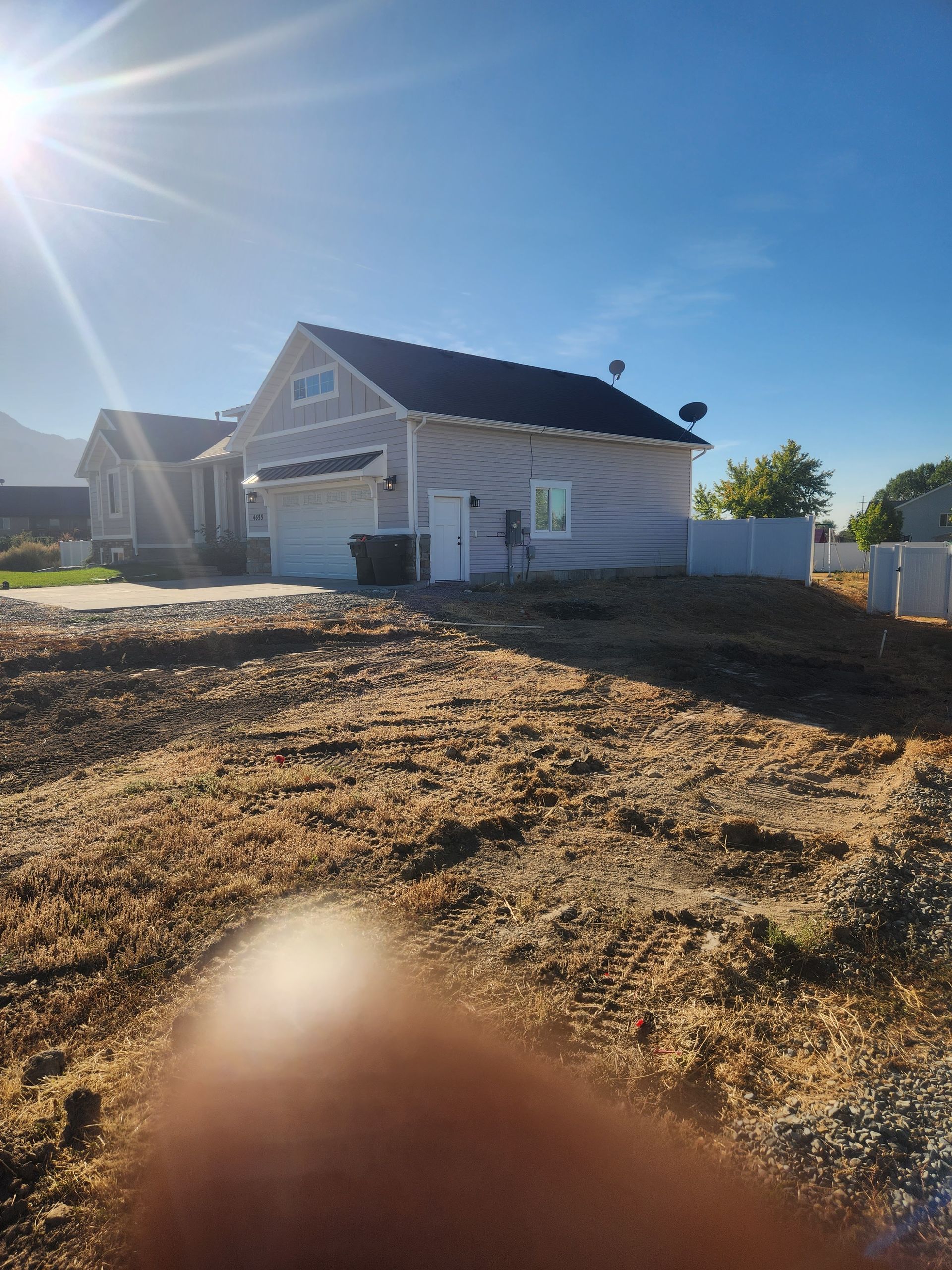 A light gray house with a black roof under a bright sun, with a bare dirt yard in front.