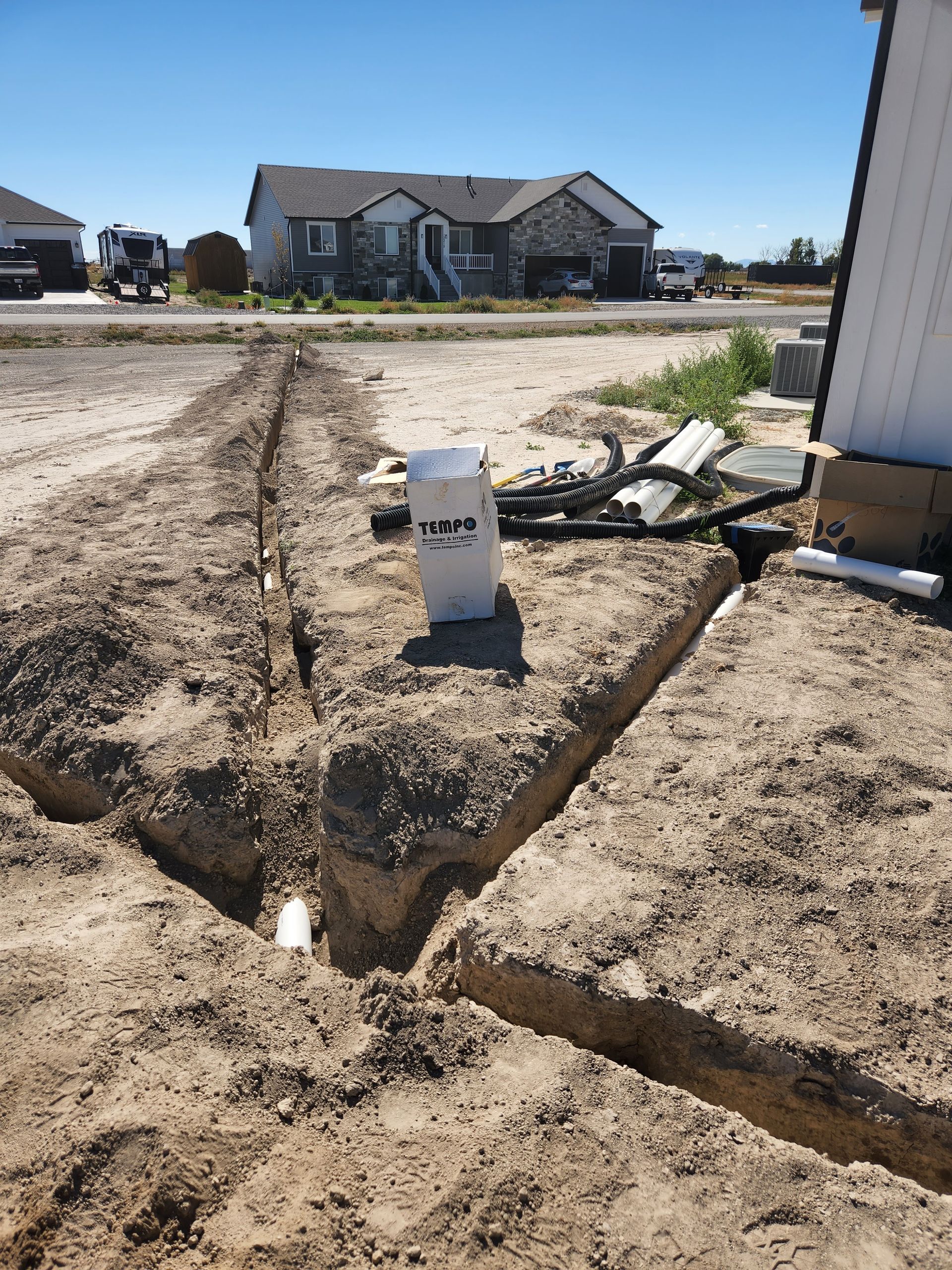 Trenches dug in dirt for underground pipes in front of a house on a sunny day.