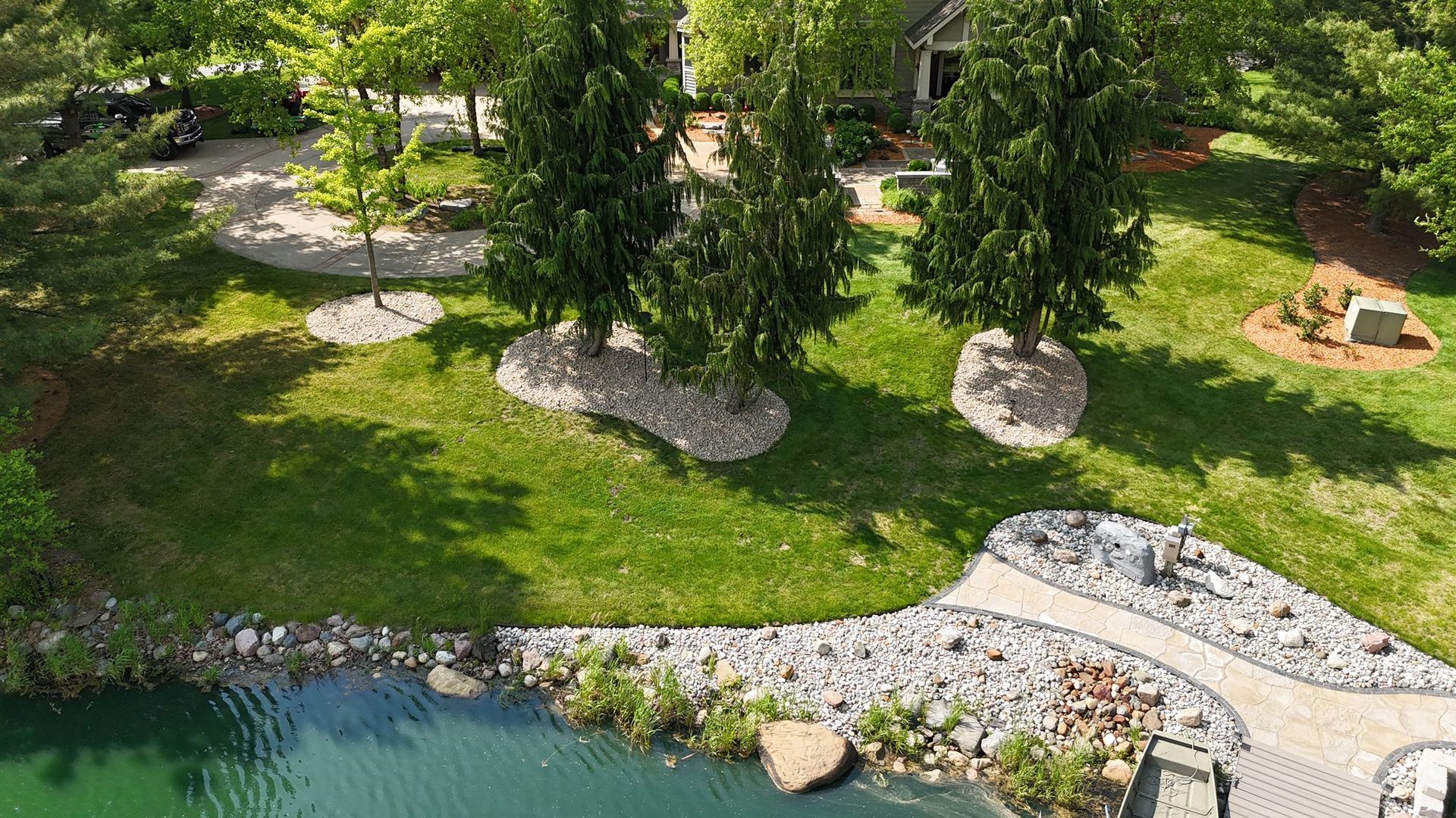 Aerial view of a landscaped yard with green grass, trees, and a stone walkway alongside a pond.