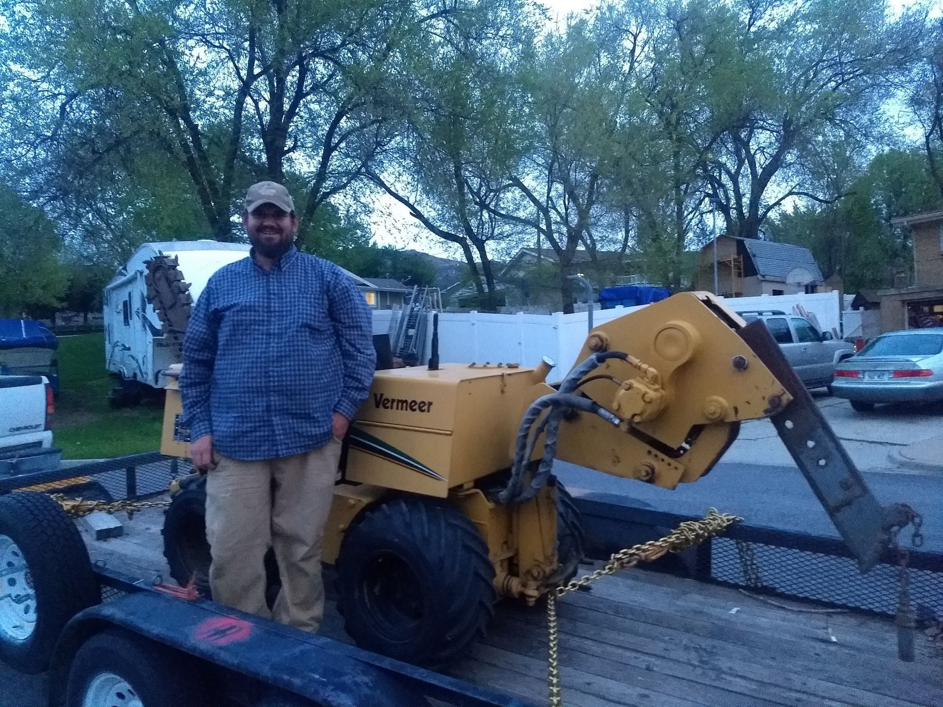Man standing next to a yellow stump grinder on a trailer, in a yard.