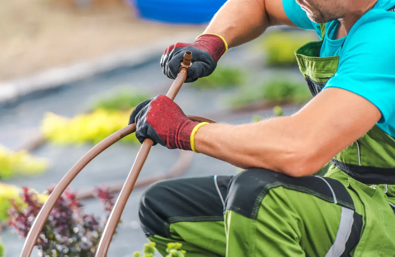 Person in green overalls and gloves connecting a brown hose outdoors.