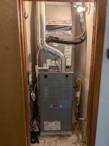 Furnace in a narrow closet with exposed ductwork and pipes, framed by wooden door jambs.