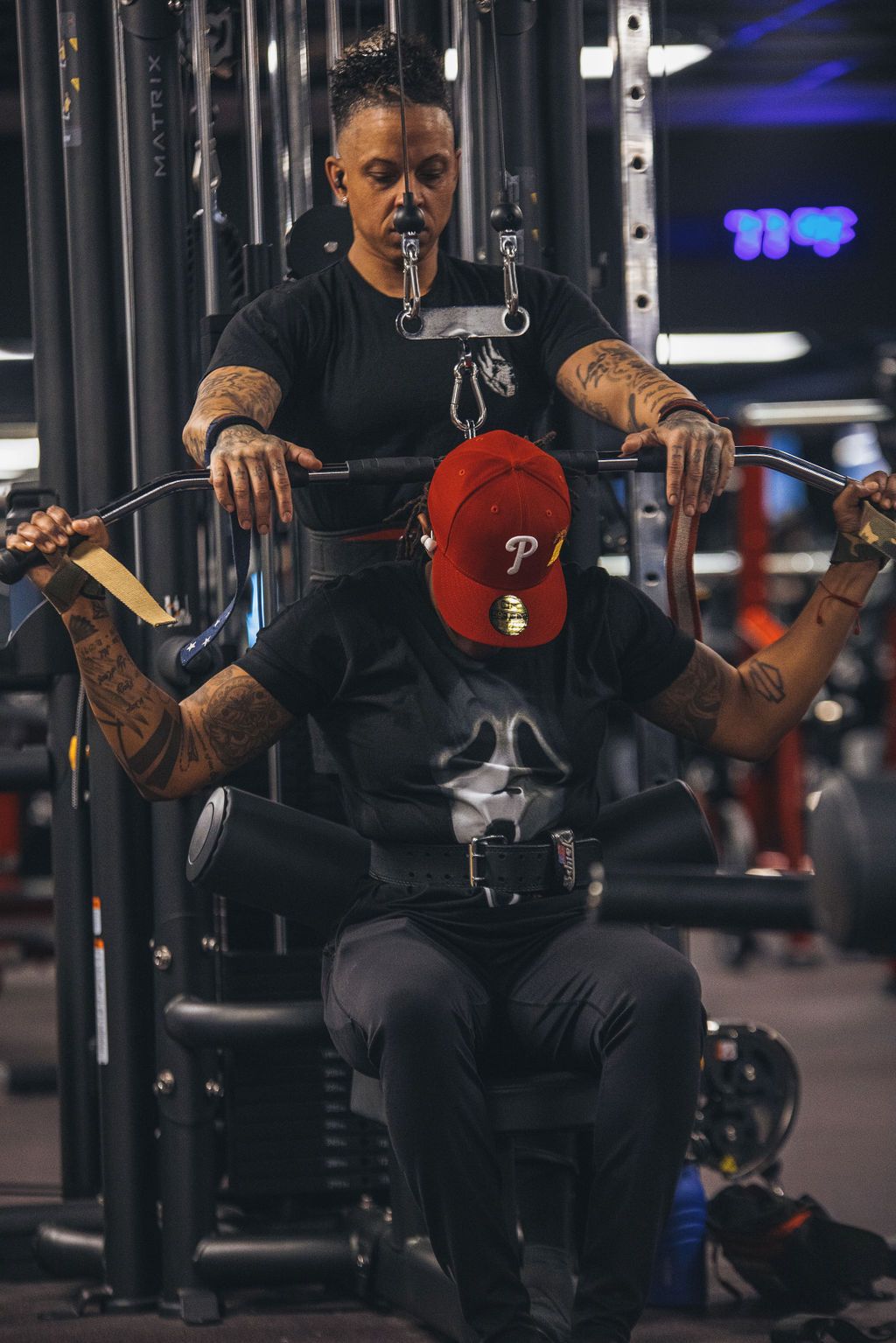 A man is sitting on a machine in a gym while a woman helps him lift a barbell.