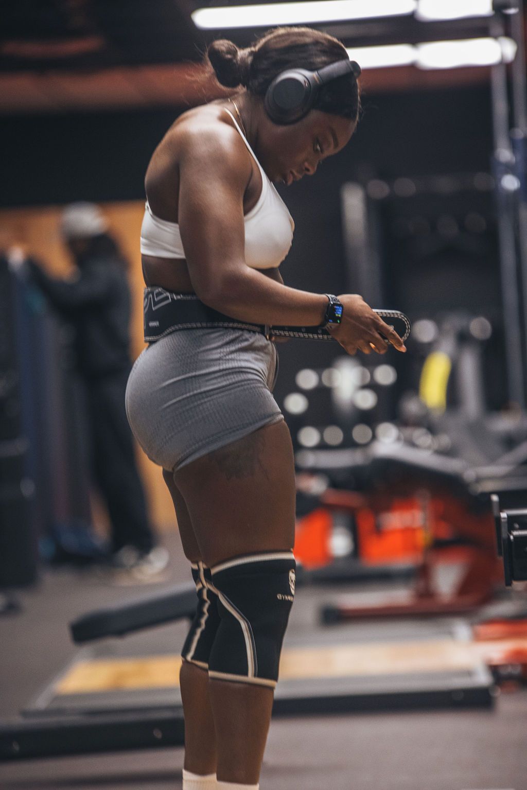 A woman wearing headphones and knee pads is standing in a gym.