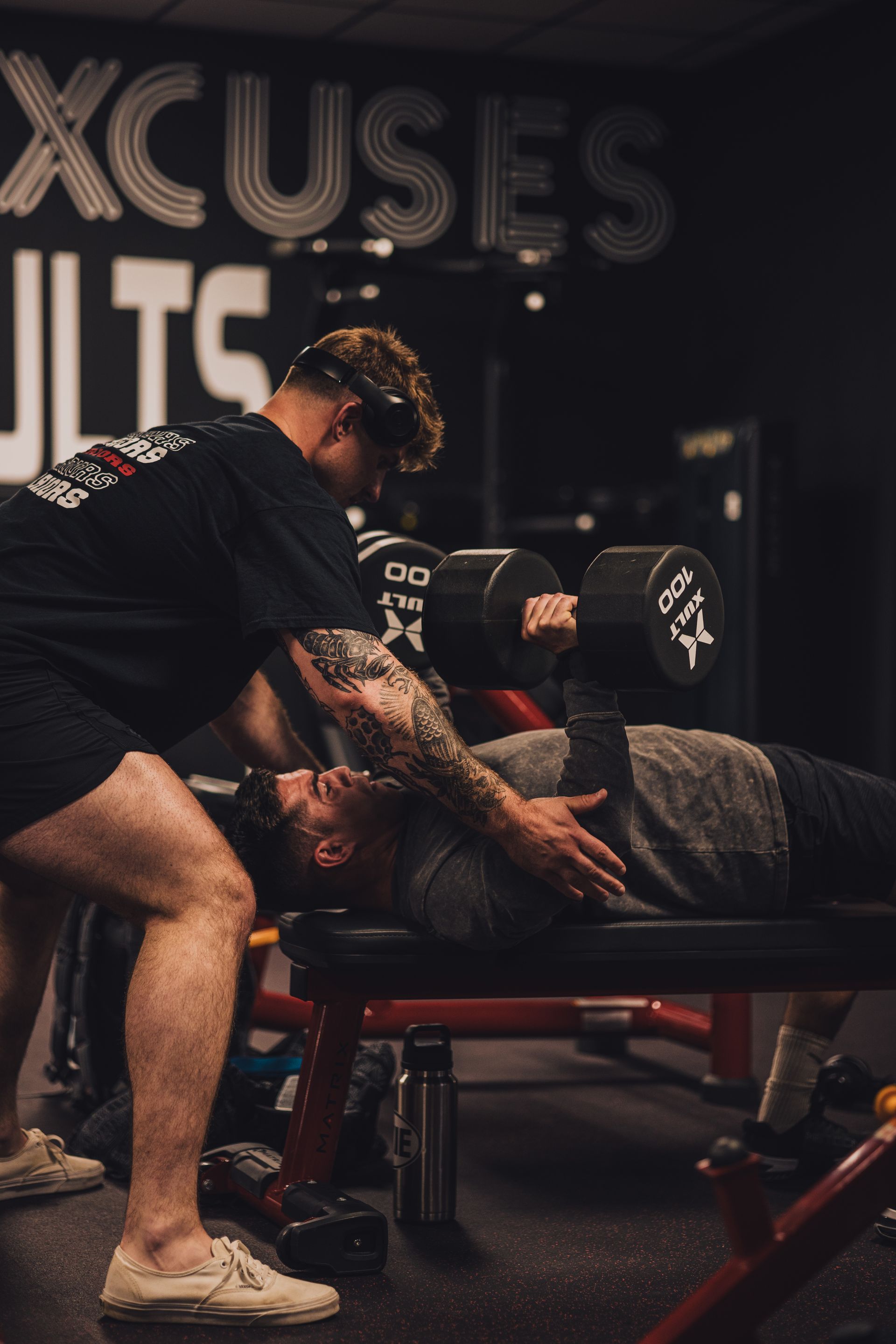 A man is helping another man lift a dumbbell in a gym.