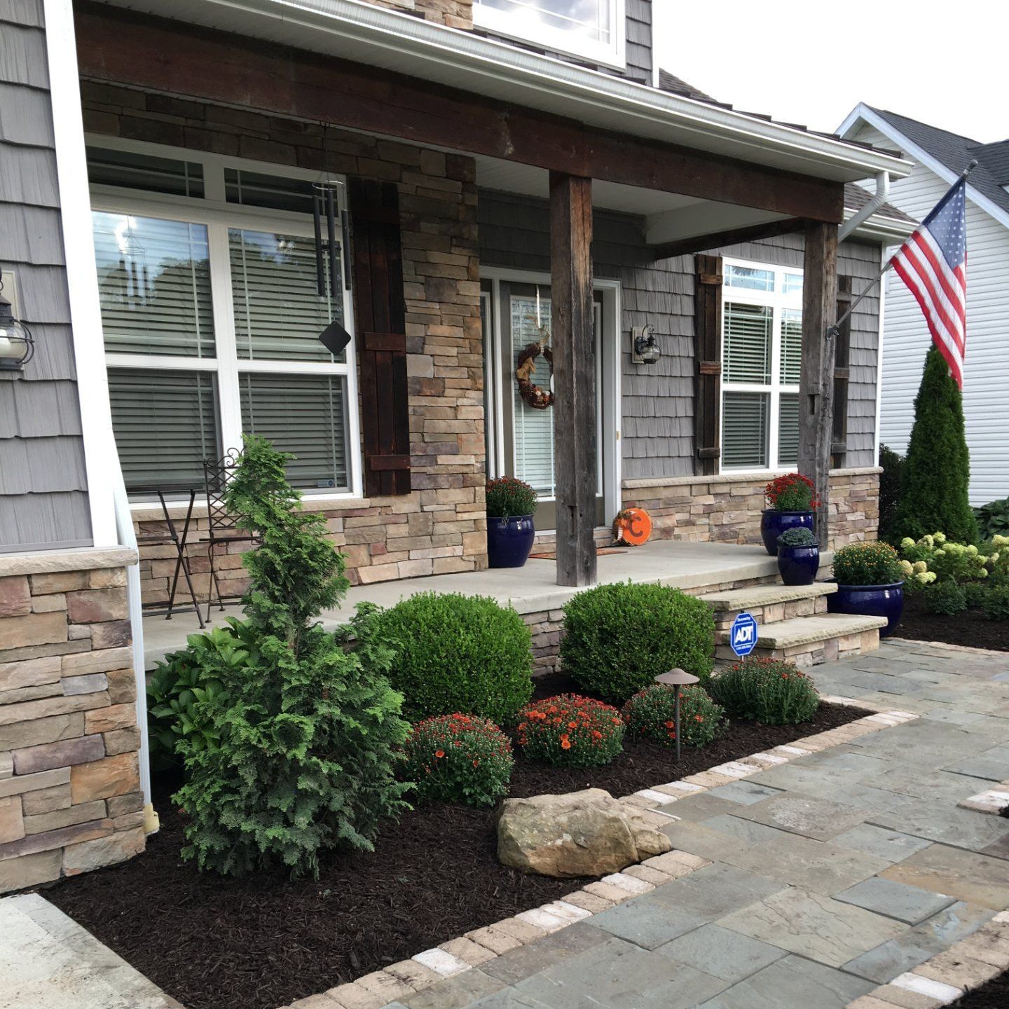 Front porch with stone and wood accents, shrubs, flower pots, walkway, and American flag.