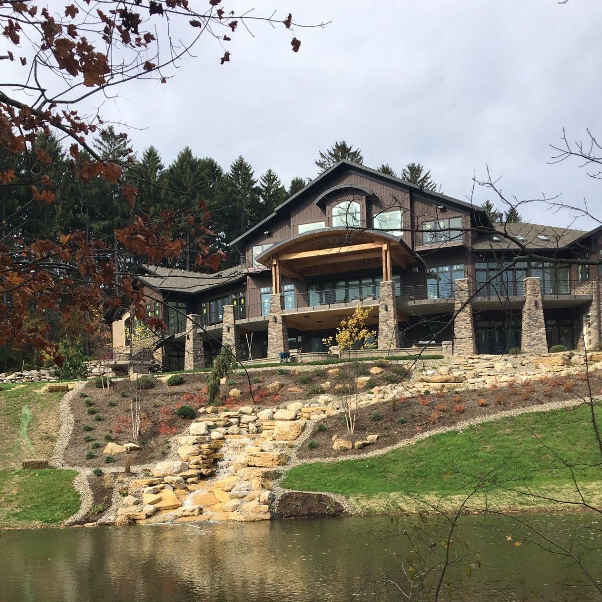 Large wood and stone house on a hill overlooking a lake; fall foliage and cloudy sky.