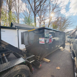 Black flatbed trailer beside a brick house on a grassy yard, with a raised rear gate.