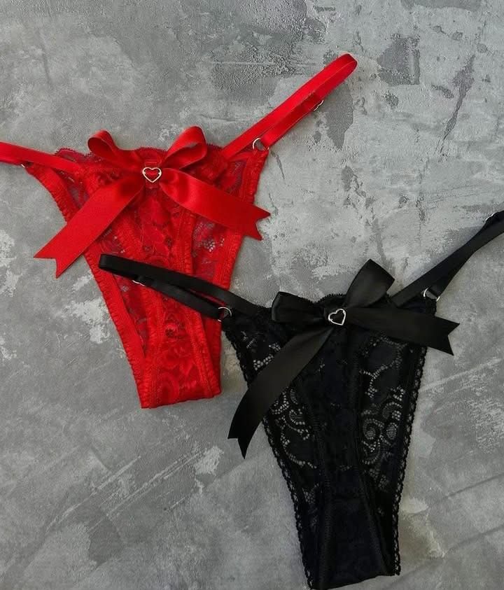 Ropa interior tipo tanga de encaje rojo y negro con lazos y detalles en forma de corazón, sobre fondo gris.