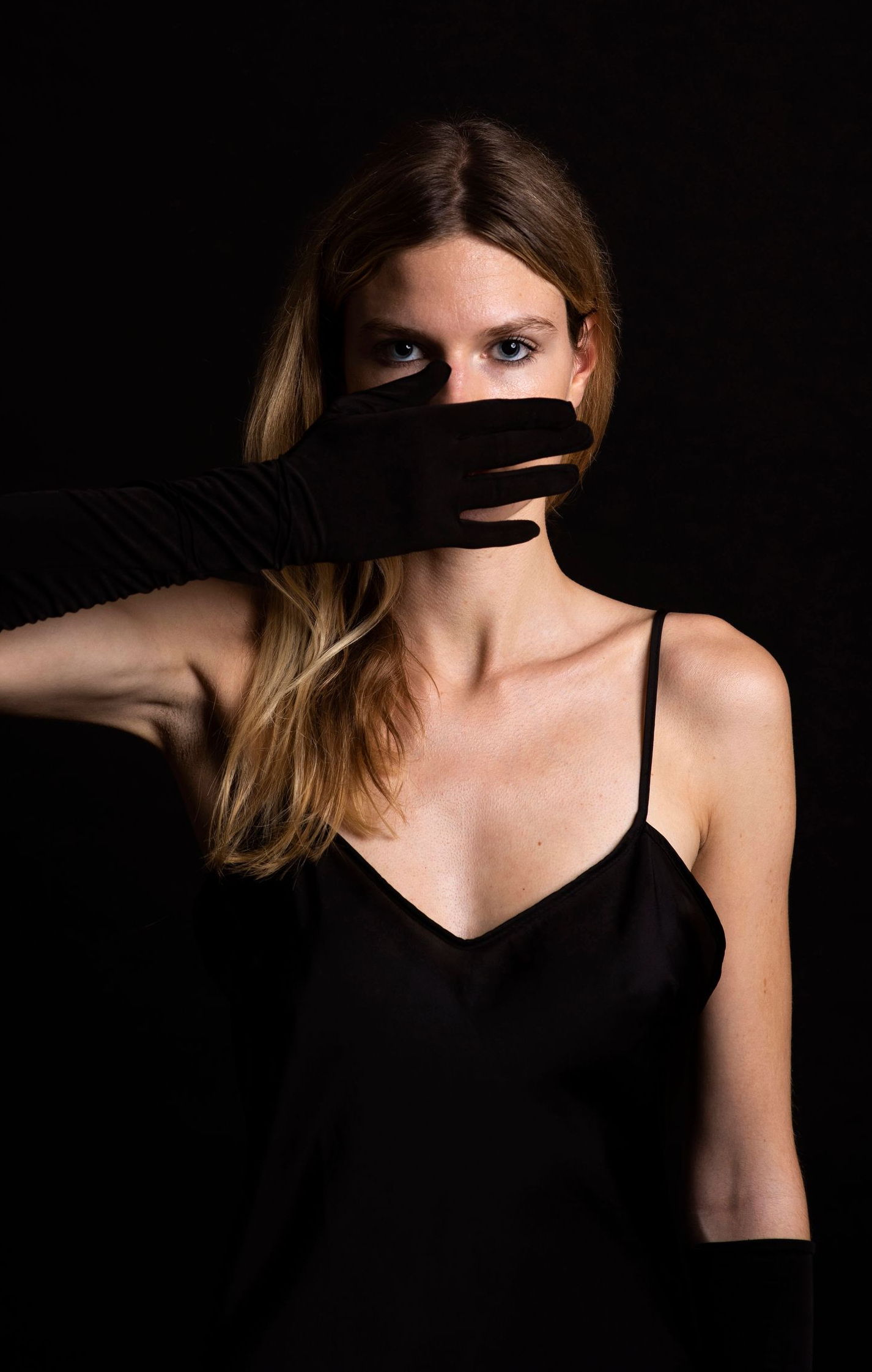 Mujer vestida de negro con un guante negro cubriéndose la boca. Fondo oscuro.