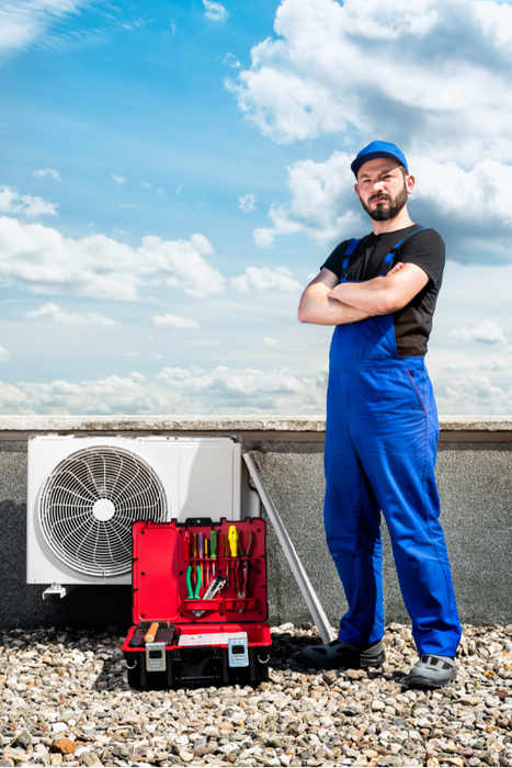 A man in blue overalls is standing on a roof next to a fan and a toolbox.