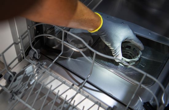 Gloved hand placing a dishwasher filter into the bottom rack of a stainless steel dishwasher