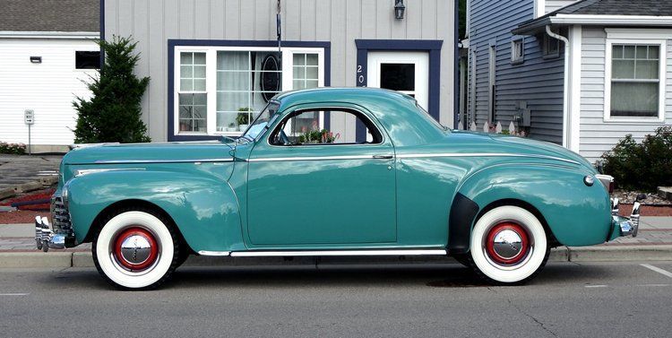 A blue car with white tires is parked in front of a house