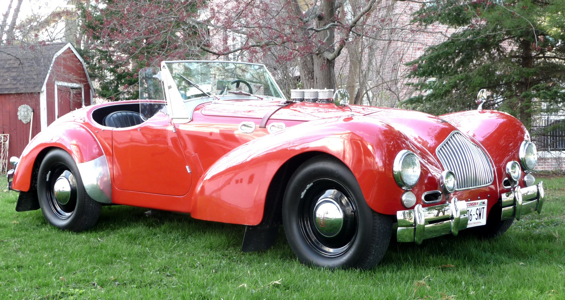 A red car is parked in the grass in front of a barn.