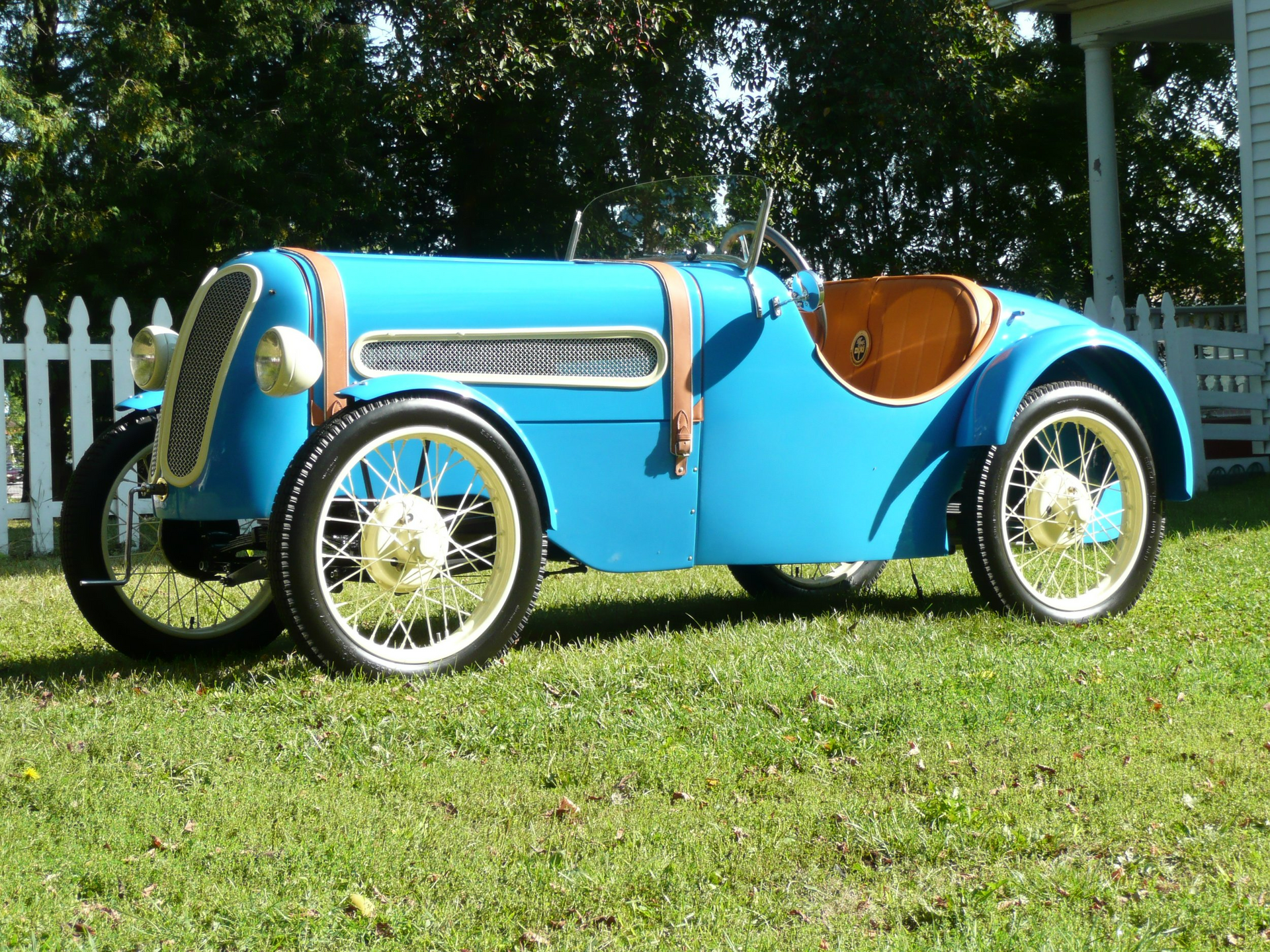 A blue car is parked in a grassy field with a white picket fence in the background