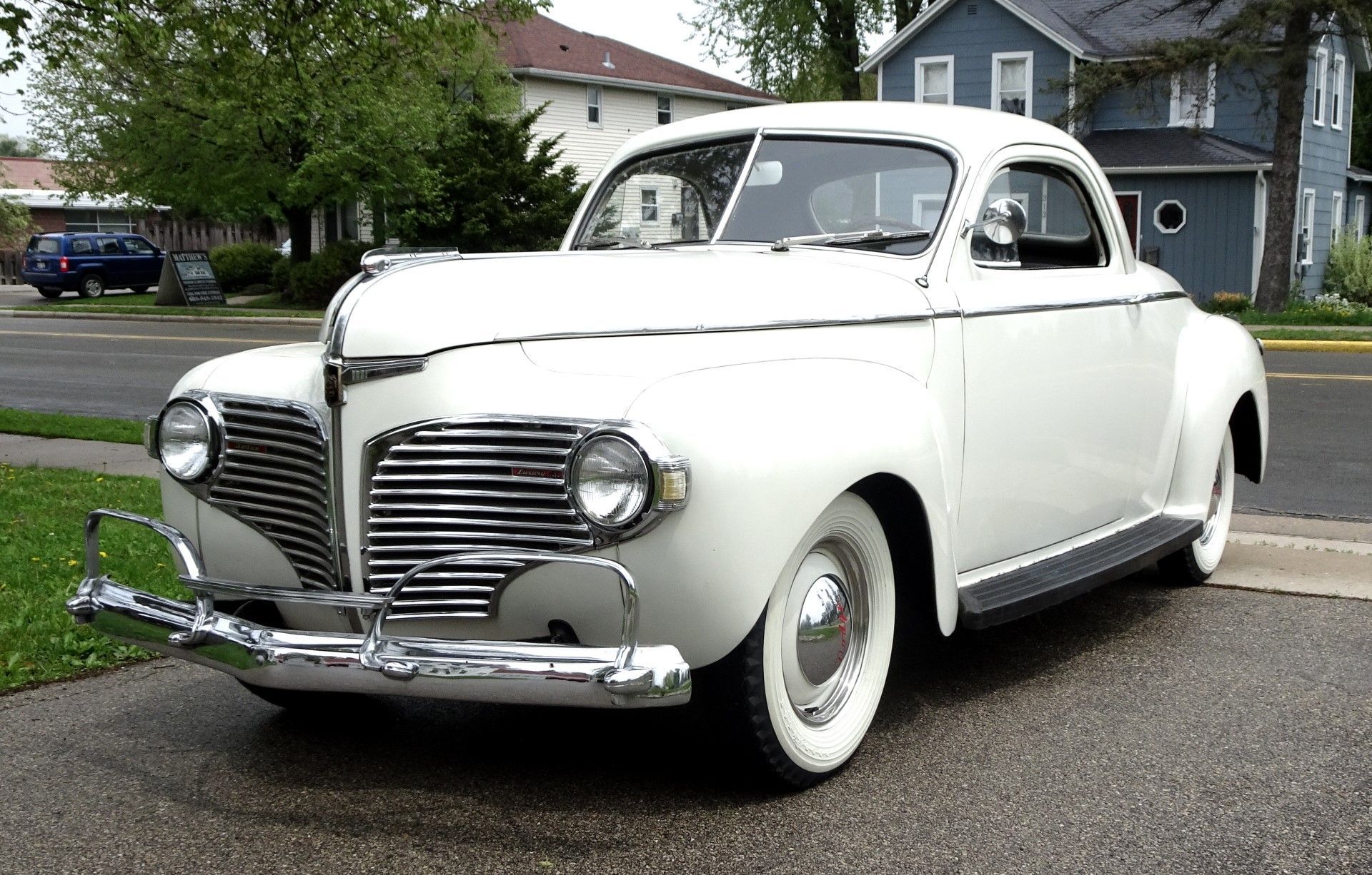 A white car is parked on the side of the road in front of a blue house.