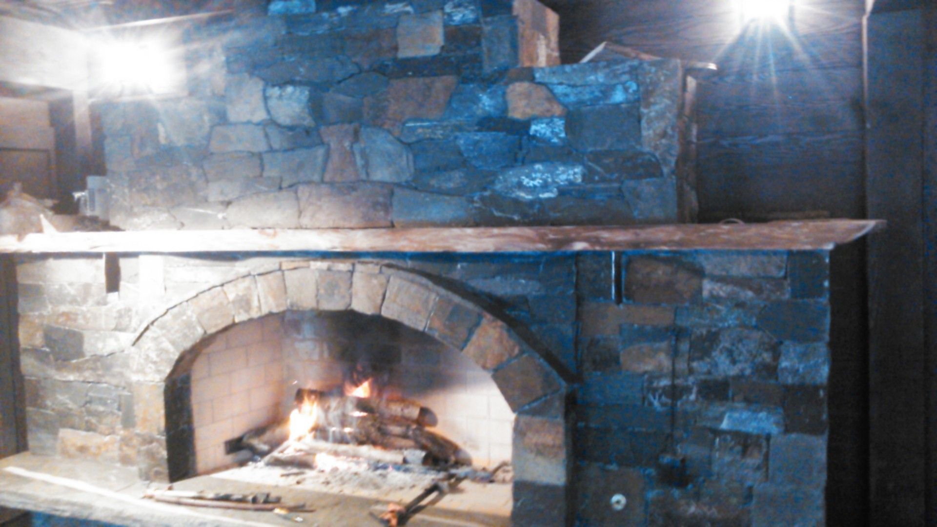 Fireplace with burning fire, wood pile, cozy chair, and brick wall.