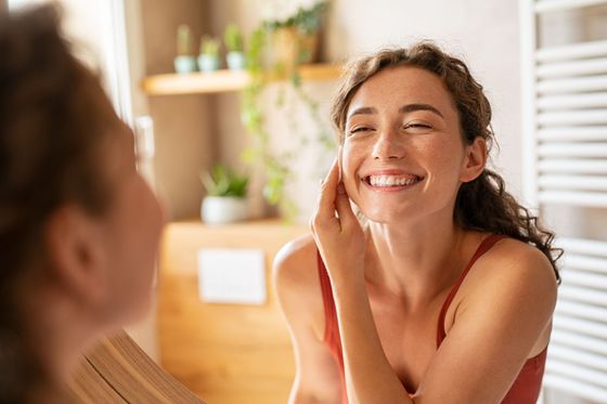 Woman in red top smiling while applying skincare in a bathroom, looking at her reflection.