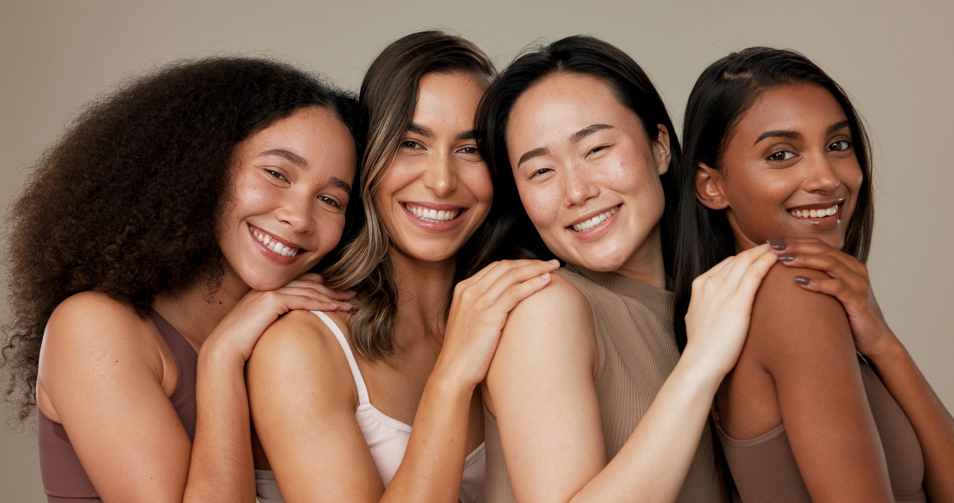 Four diverse women smiling and embracing, skin tones varying, neutral background.