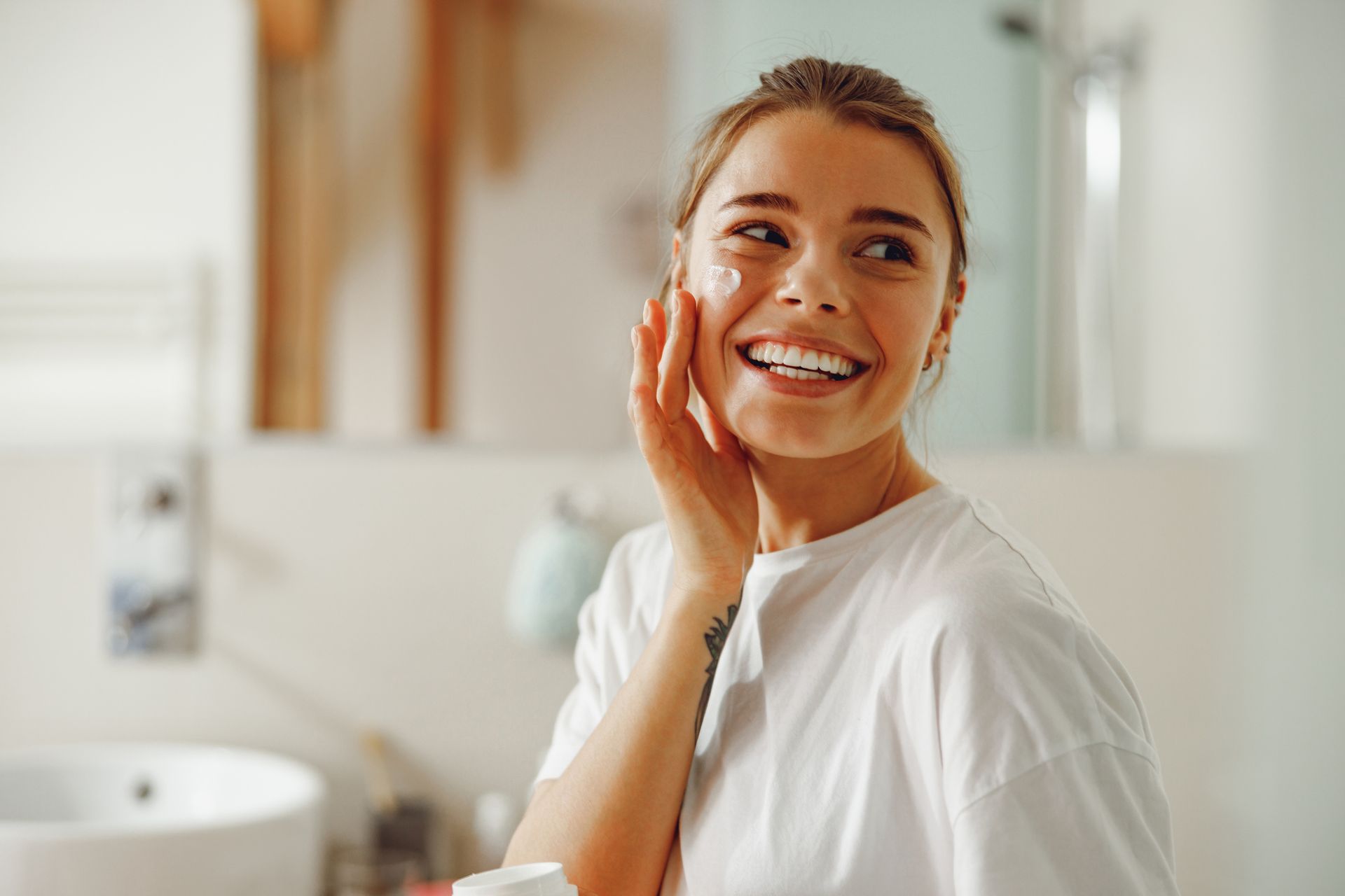 A woman is smiling while applying cream to her face in a bathroom.