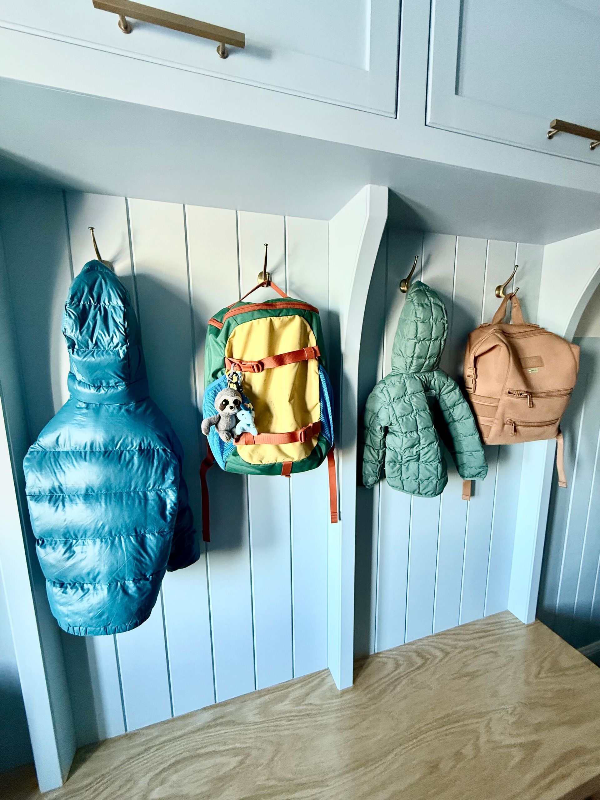 Three backpacks and jackets are hanging on hooks on a wall.