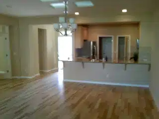An empty living room with hardwood floors and a kitchen in the background.