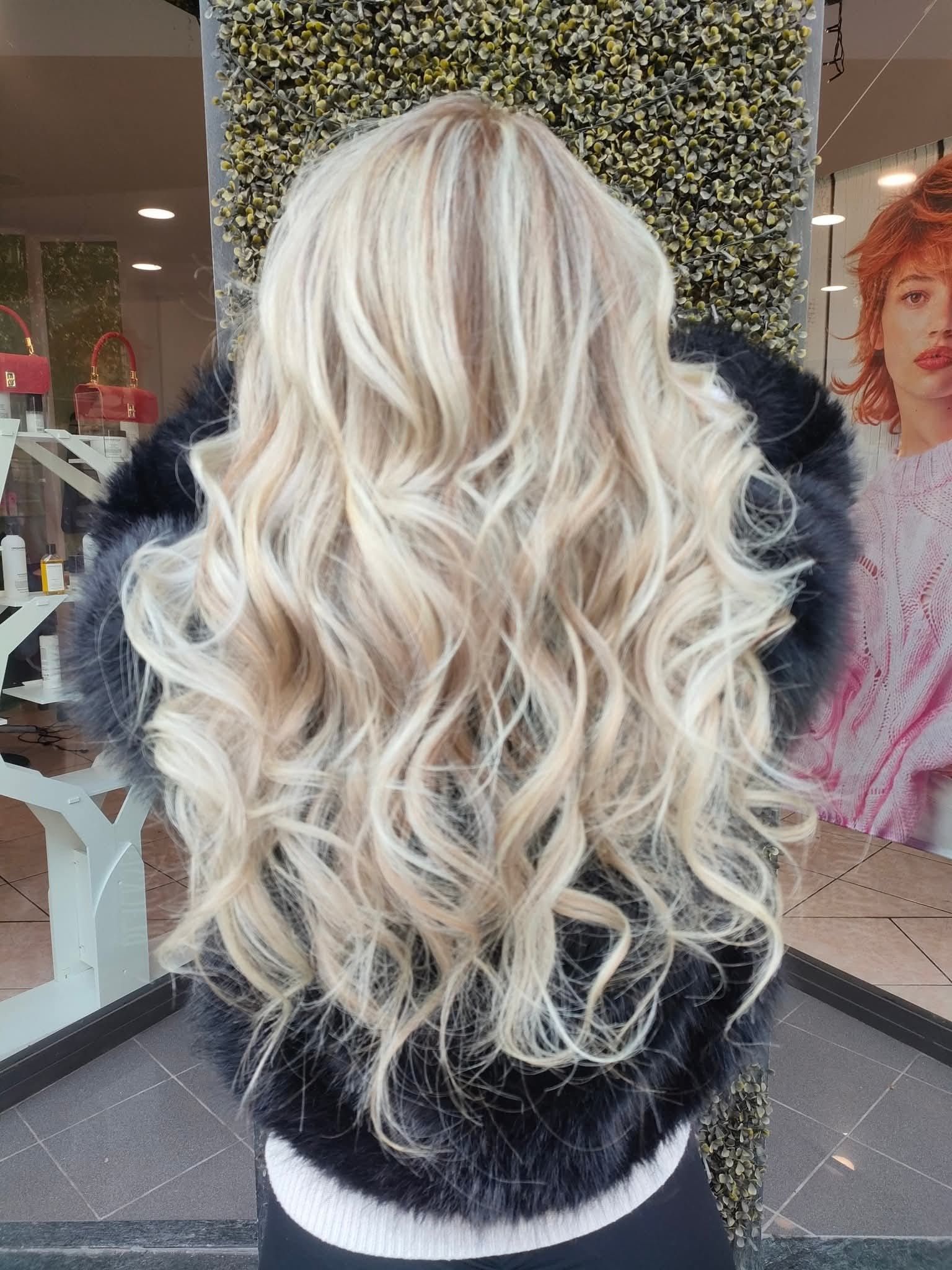 Una persona con lunghi capelli mossi color biondo platino, che indossa una giacca di pelliccia sintetica nera, davanti alla vetrina di un negozio.