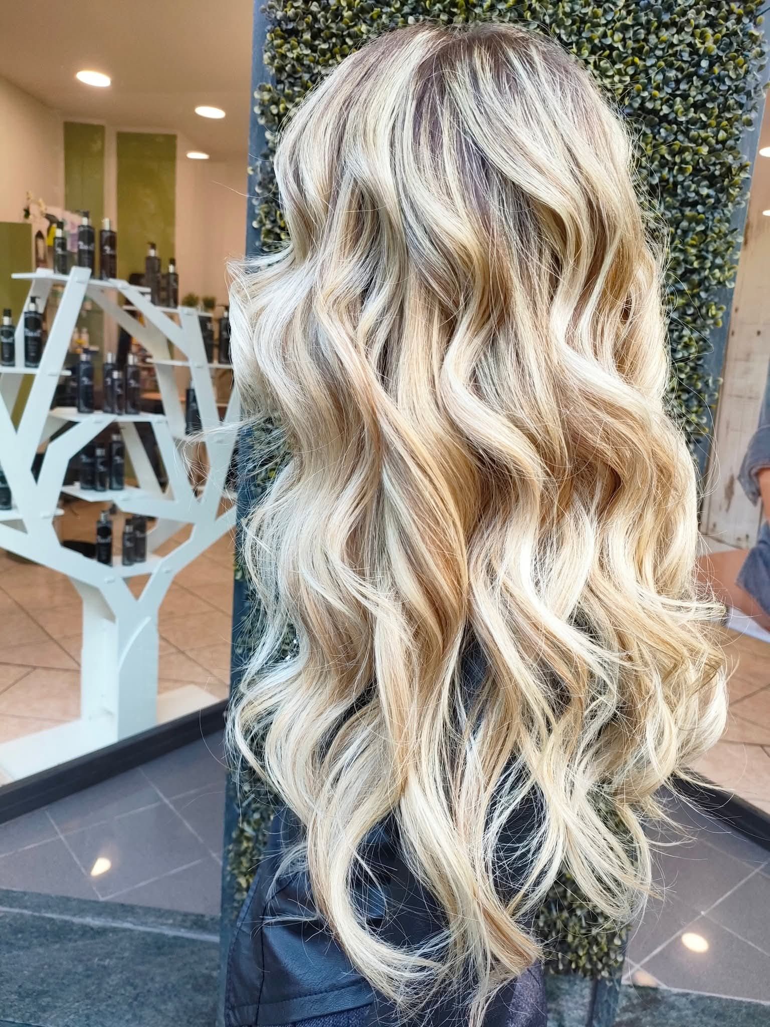 Lunghi capelli biondi ondulati con riflessi multi-tono, acconciati in un salone con una mensola decorativa a forma di albero sullo sfondo.