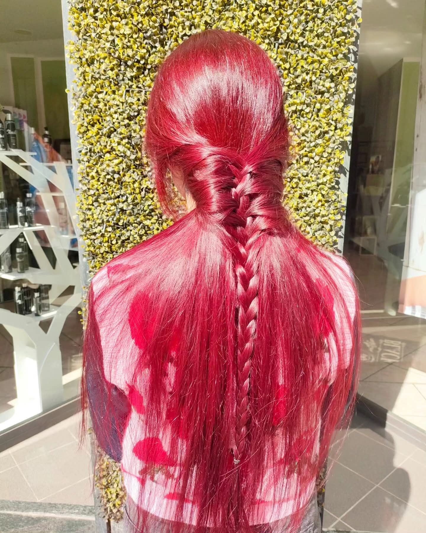 Una persona con i capelli rosso acceso, raccolti in una mezza coda intrecciata, è in piedi contro un muro ricoperto di edera verde.