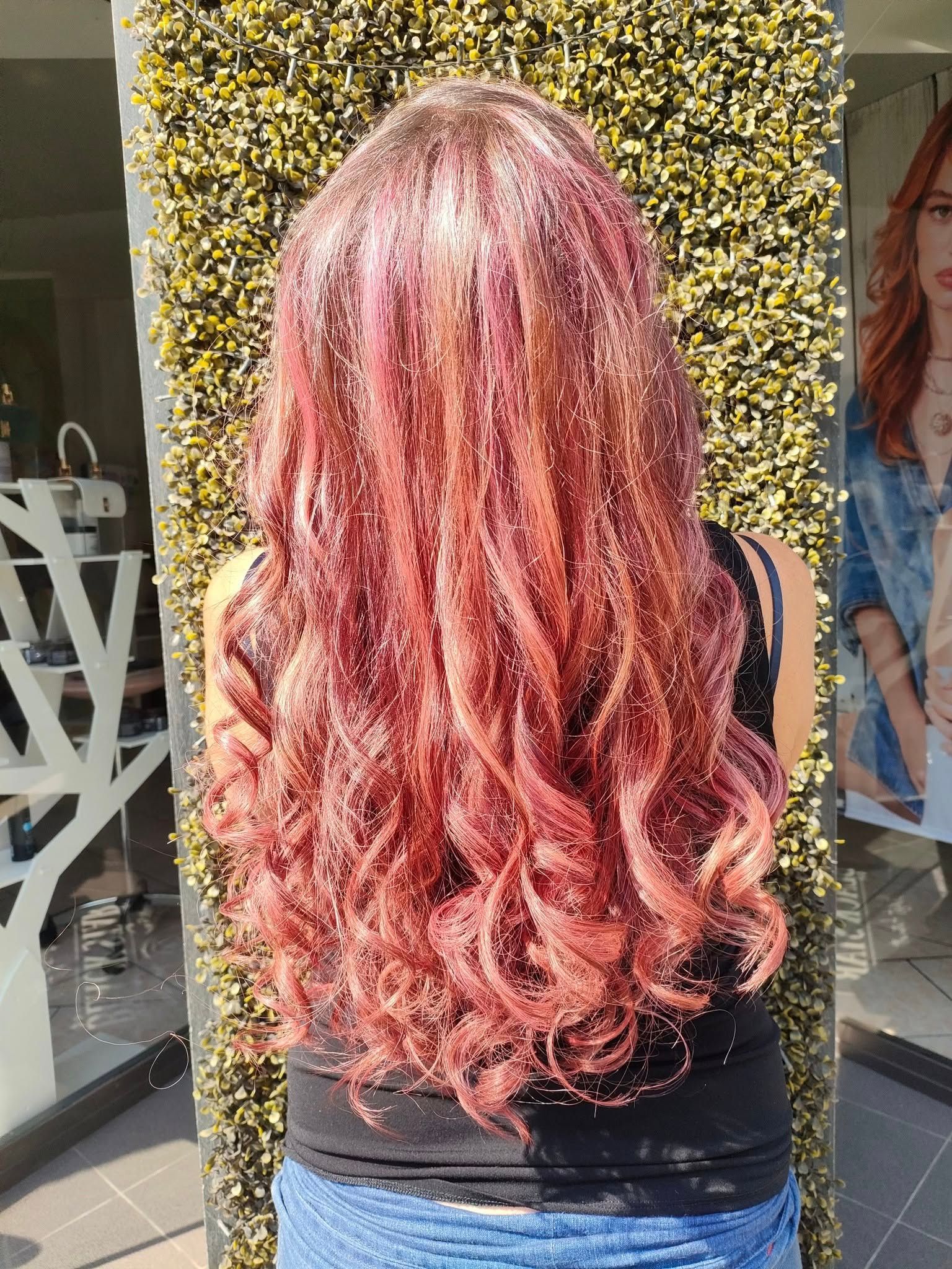 Vista posteriore di lunghi capelli ondulati in una vibrante tonalità oro rosa e rosa, portati sciolti contro un muro verde.