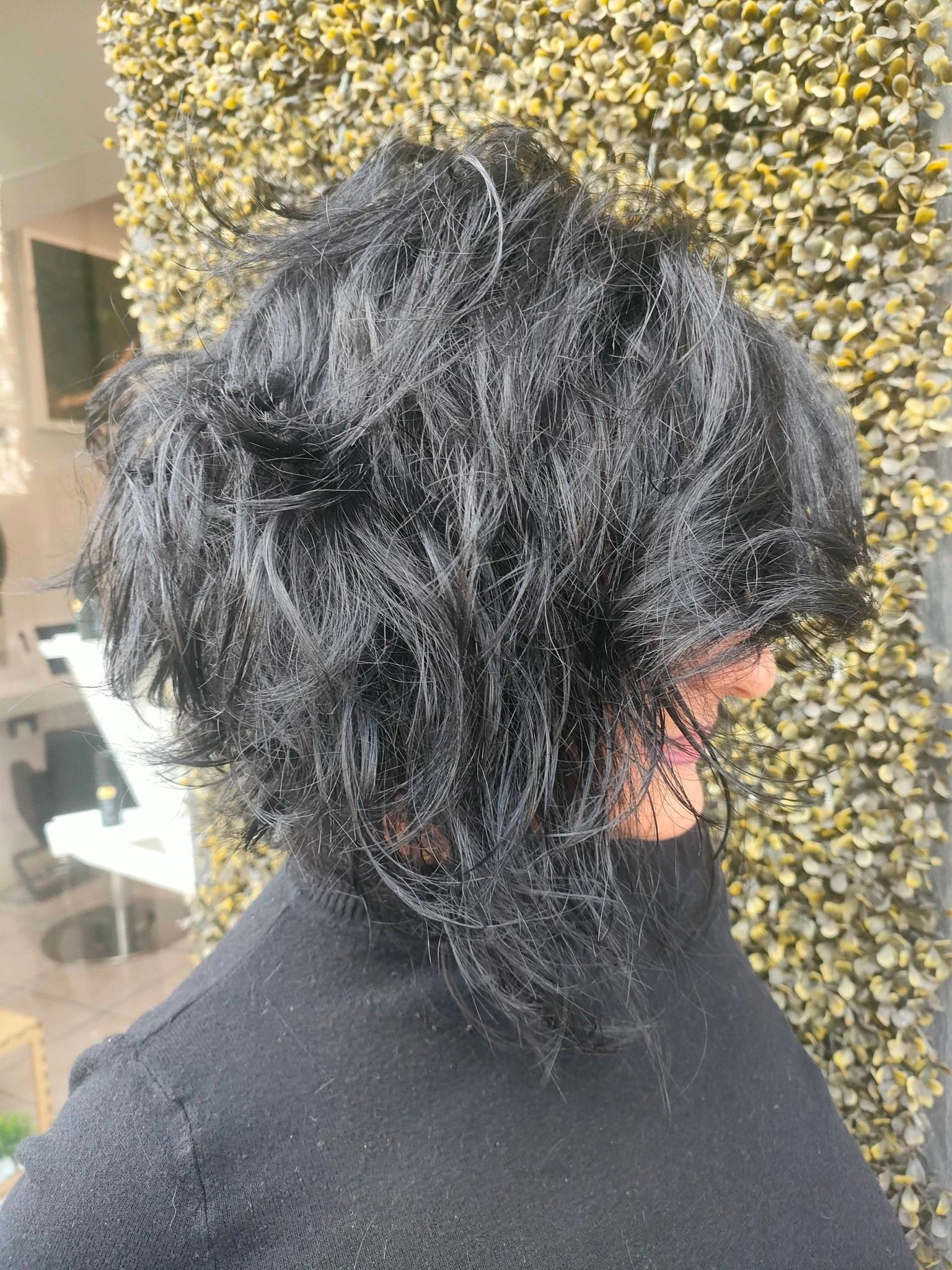 Una persona con folti capelli scuri e ondulati, tagliati a caschetto scalato, vista di profilo, in piedi contro un muro ricoperto di vegetazione verde.