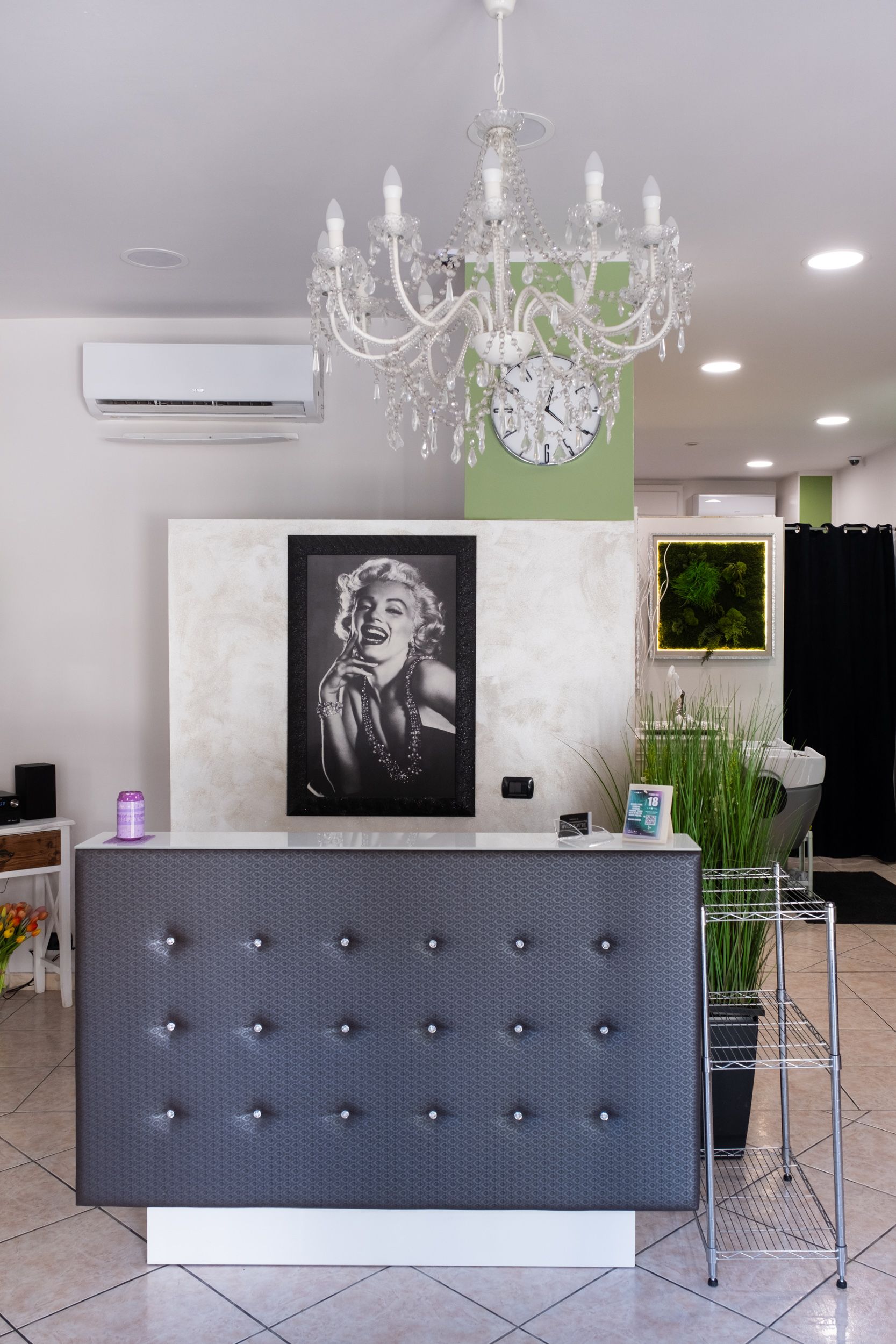 In una stanza luminosa, il bancone della reception di un salone di bellezza con un frontale grigio trapuntato, un ritratto di Marilyn Monroe e un lampadario di cristallo.