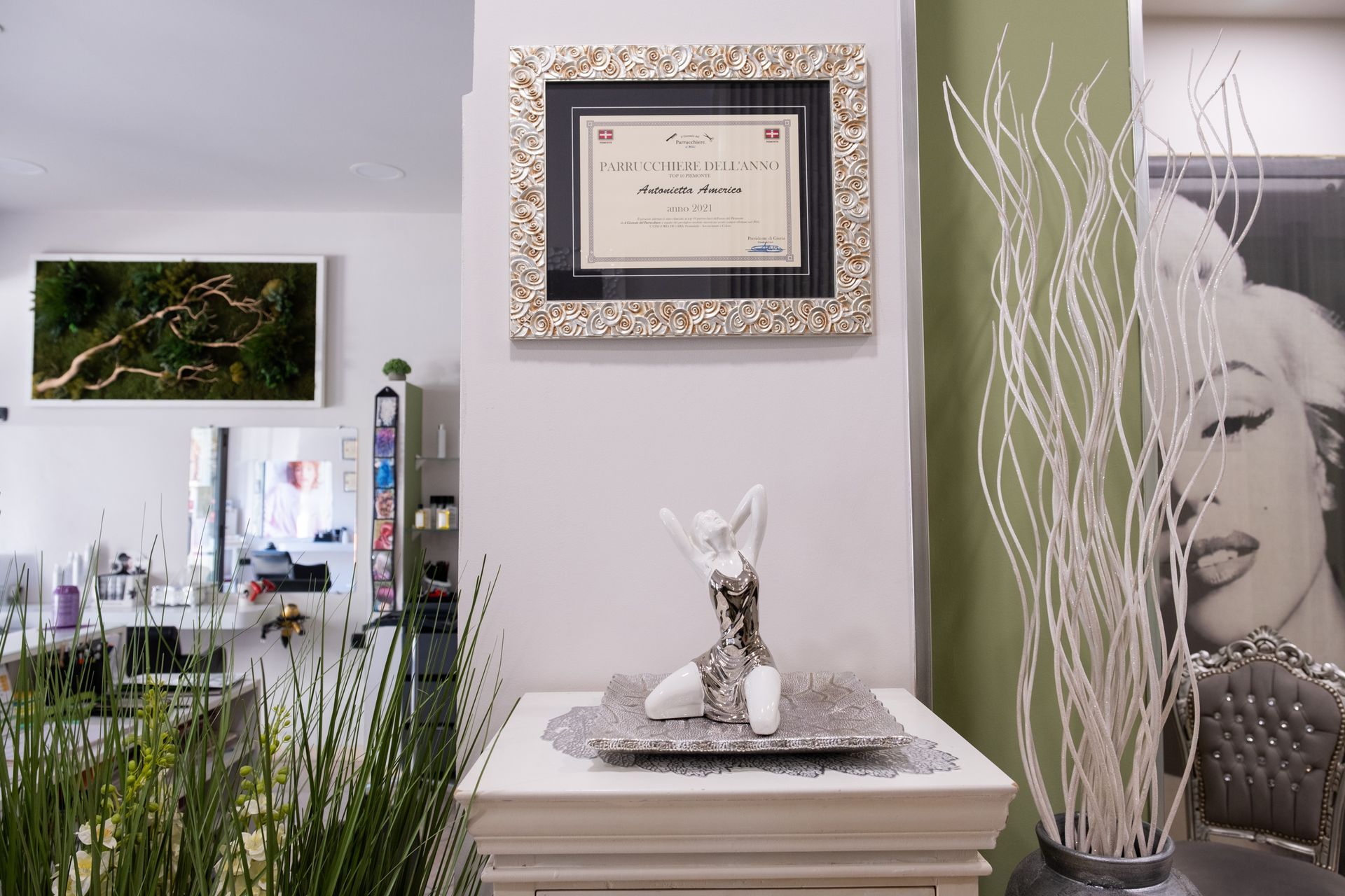 In una stanza con un certificato incorniciato, dettagli verdi alle pareti e alti rami bianchi, un piedistallo bianco sorregge una statuetta decorativa.