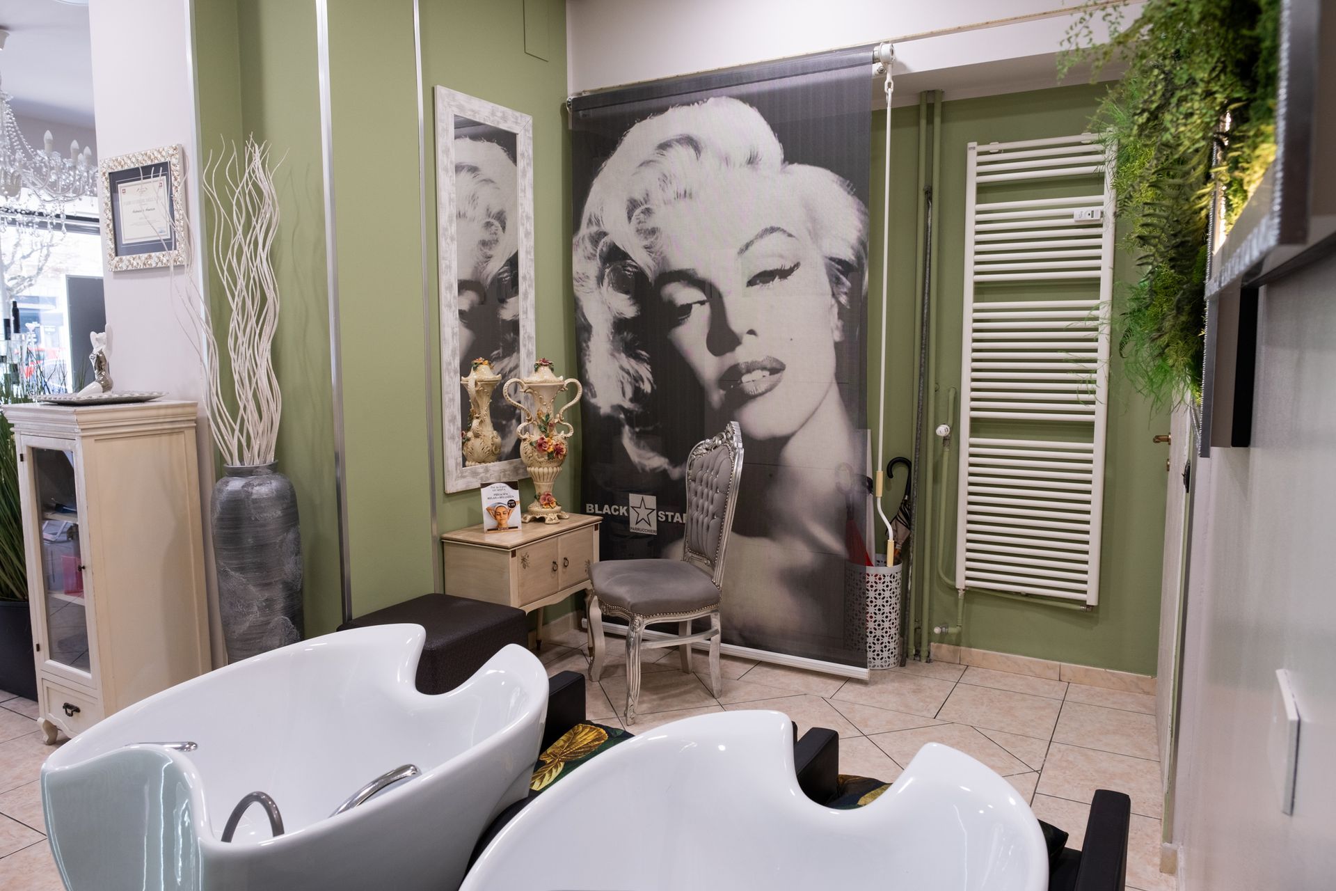 Interno di un salone da parrucchiere con due lavatesta bianchi in primo piano, pareti verde oliva e un ritratto di Marilyn Monroe.