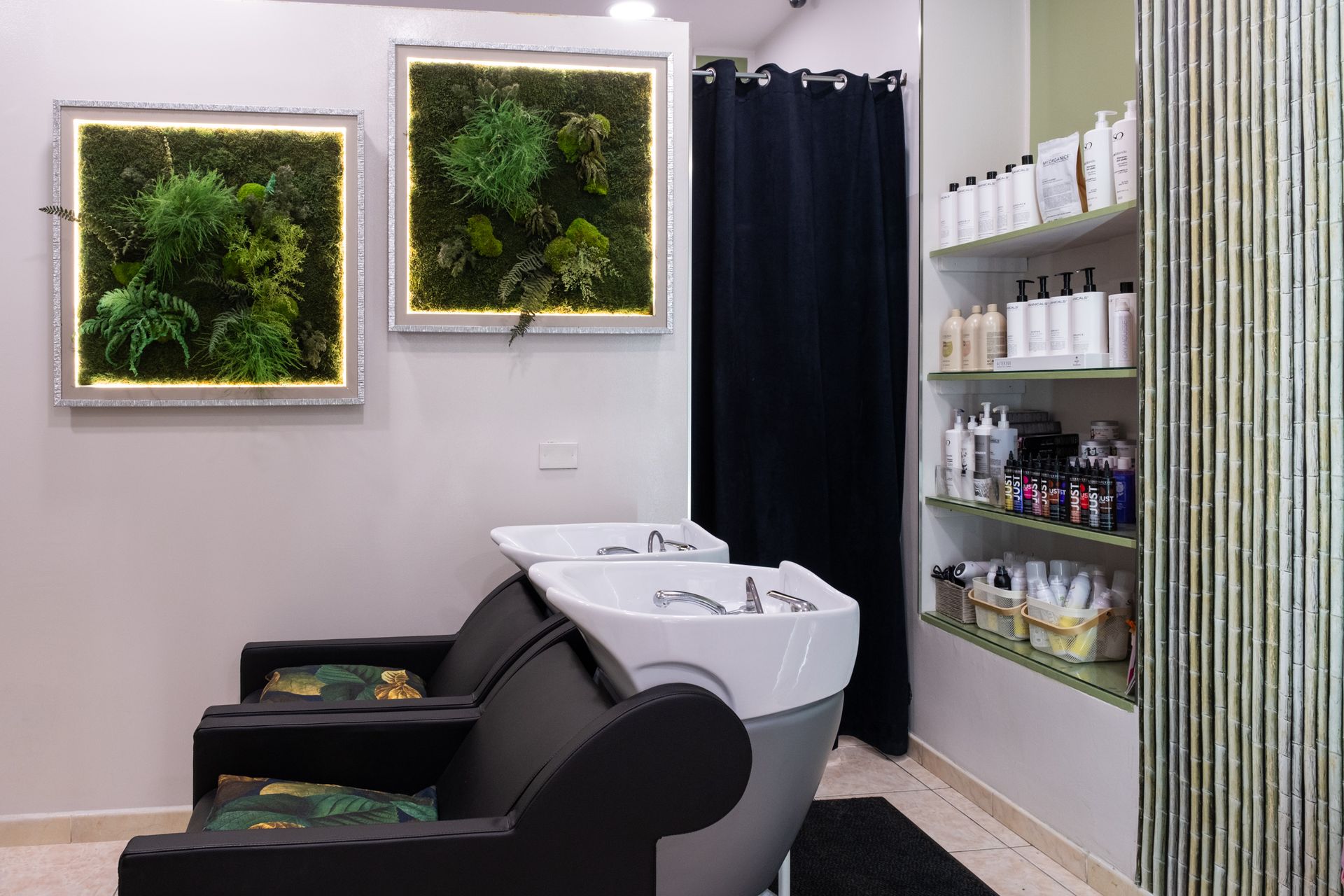 Due postazioni per il lavaggio dei capelli con sedie nere, disposte di fronte a due opere d'arte murali realizzate con muschio, accanto a scaffali riforniti di prodotti per capelli.