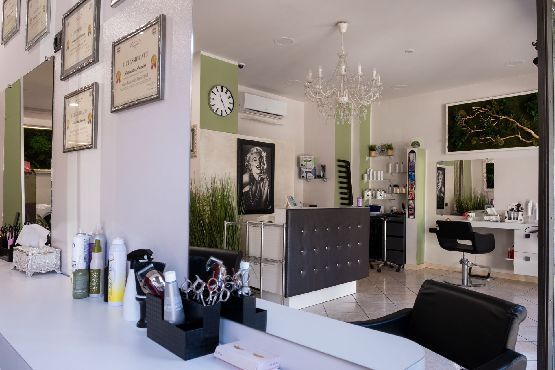 Vista interna di un luminoso salone da parrucchiere, con poltrone per lo styling, una reception, un grande lampadario e certificati appesi alle pareti.
