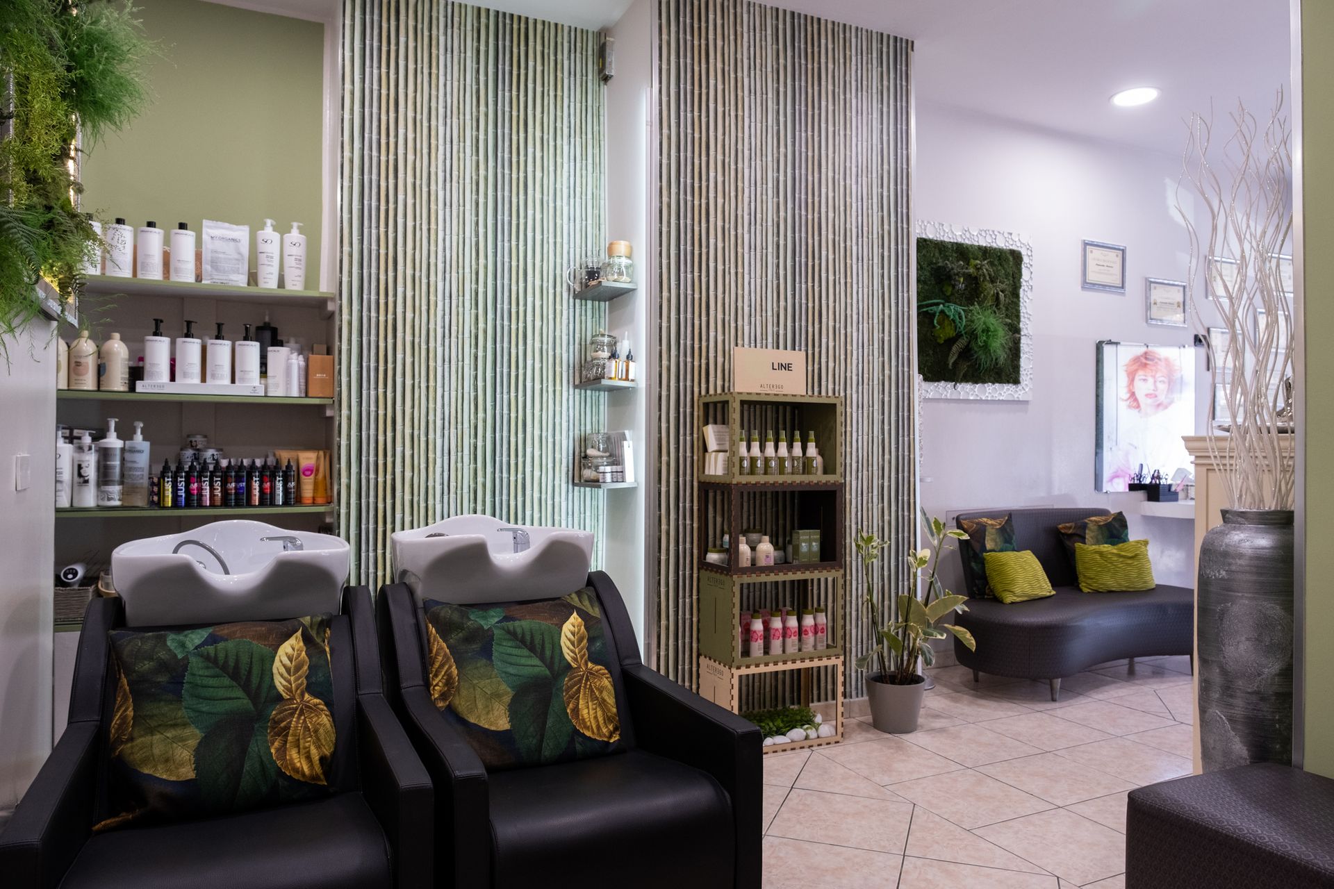 L'interno di un salone da parrucchiere con due poltrone nere per lo shampoo con cuscini a tema tropicale, scaffali con prodotti e una zona d'attesa.