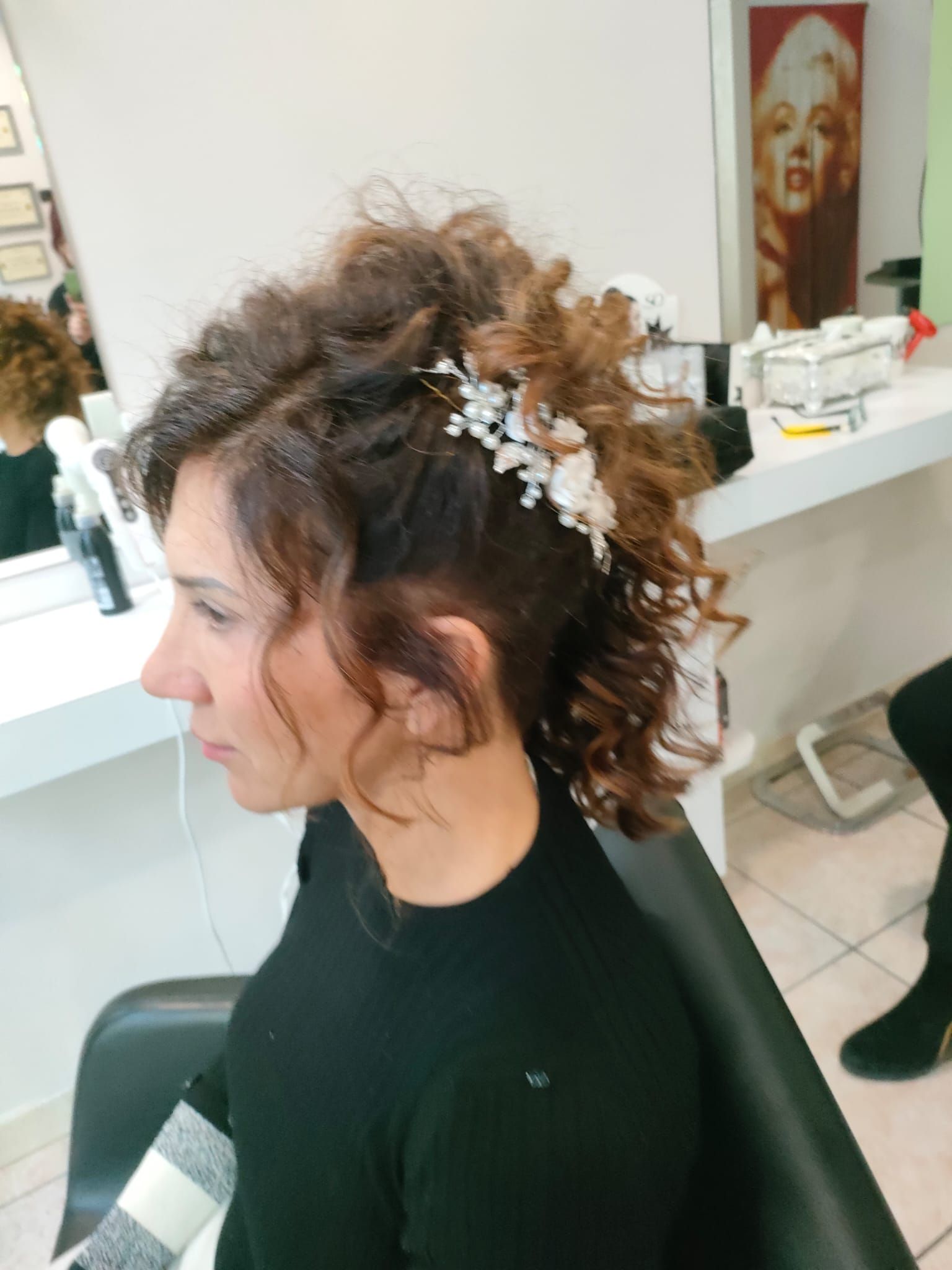 Una donna con i capelli castani ricci, raccolti in una mezza coda con un accessorio floreale, è seduta su una poltrona da parrucchiere.